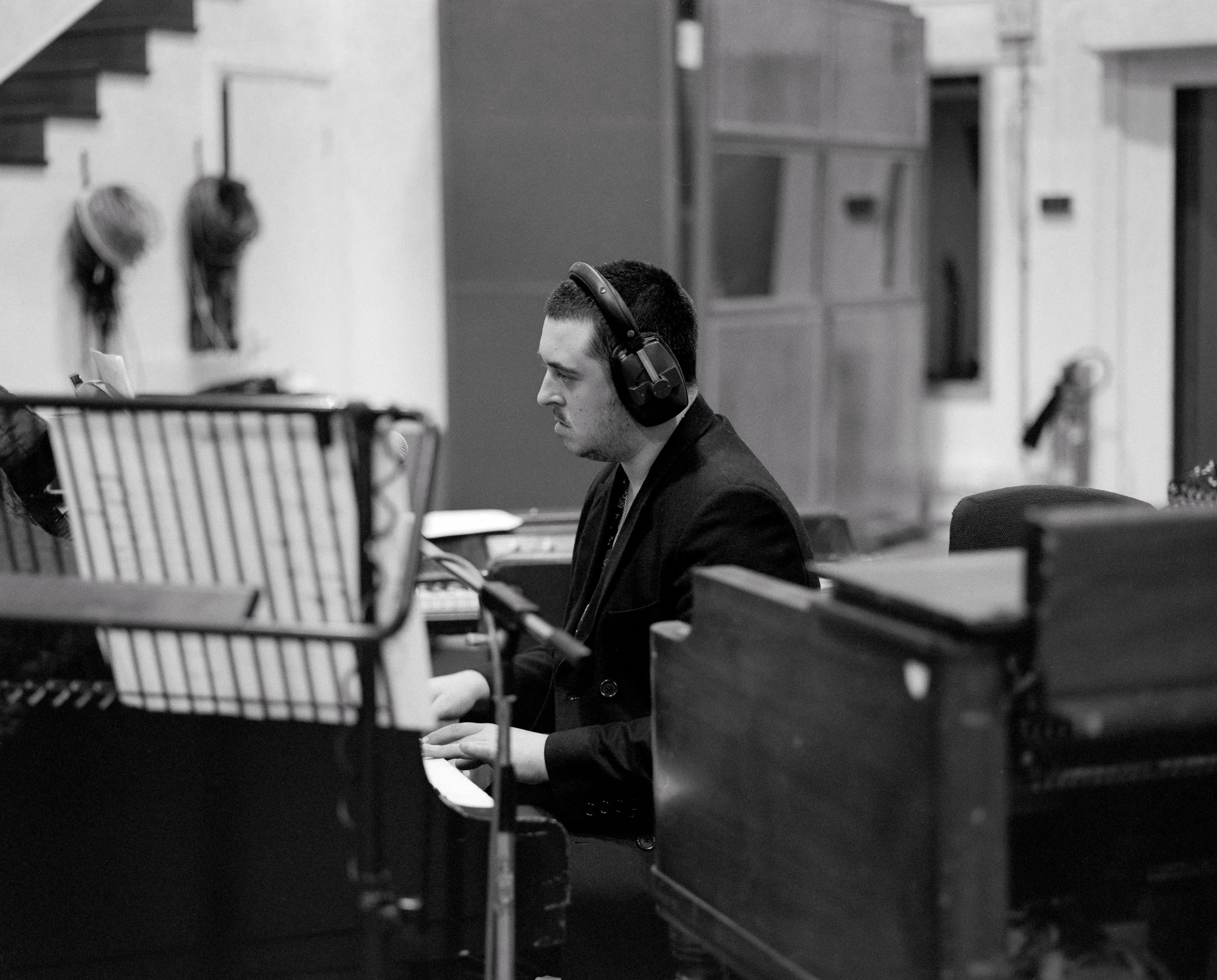 A young man with short dark hair and headphones playing a piano in a music studio, with music equipment and shelves in the background.