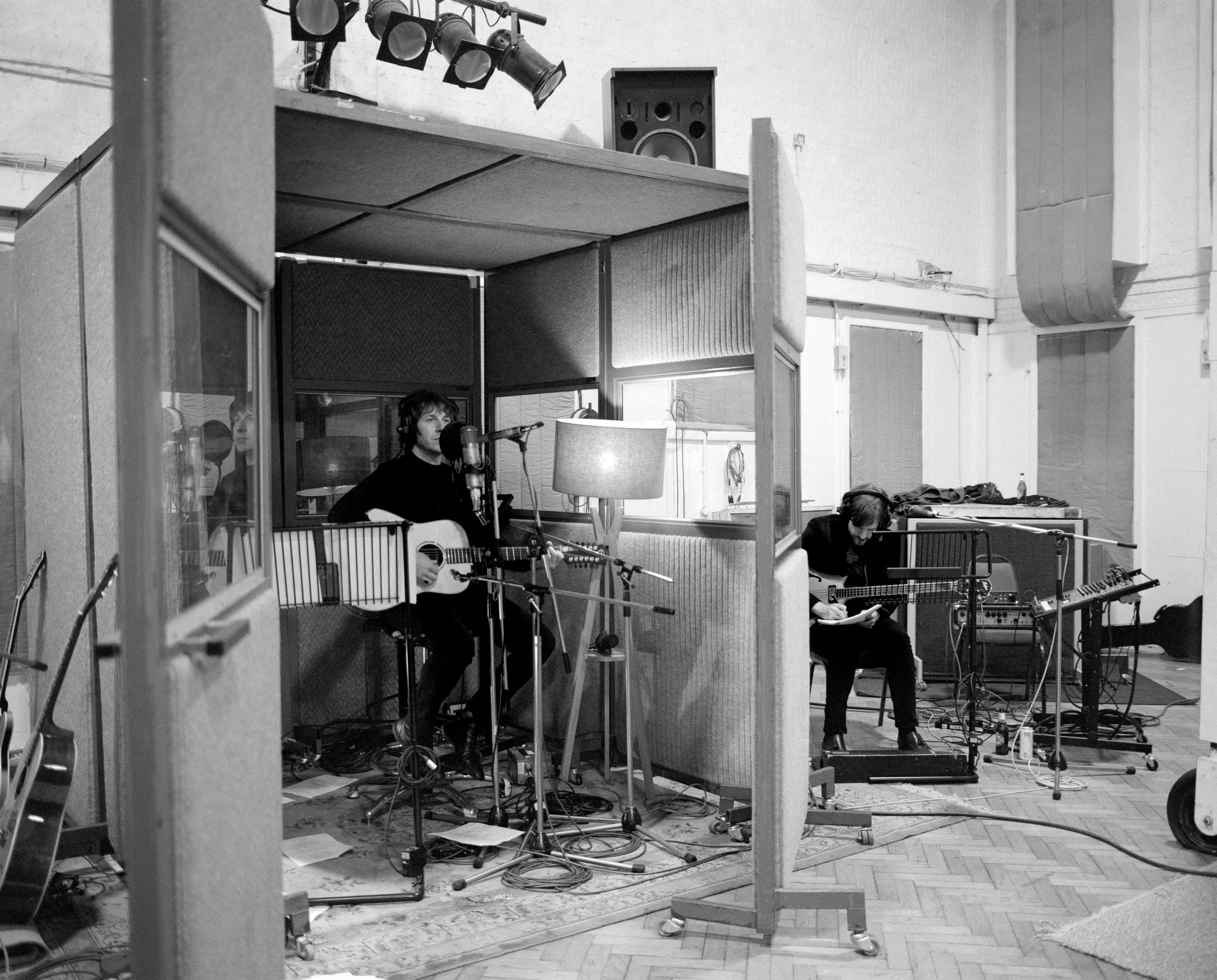 Two musicians recording in a studio, one playing an acoustic guitar and singing into a microphone, the other playing an electric guitar with a music stand nearby.