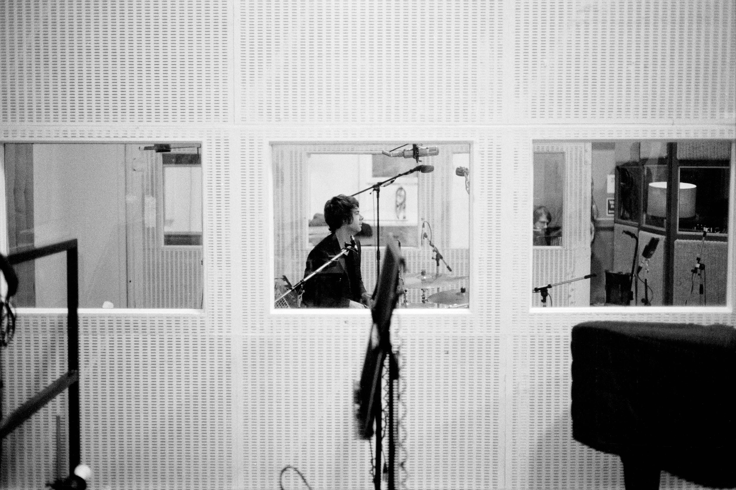 Black and white photo of a man inside a recording studio, seen through a window. The man is sitting at a drum set with a microphone above him, recording music. The studio has soundproofed walls and various audio equipment.