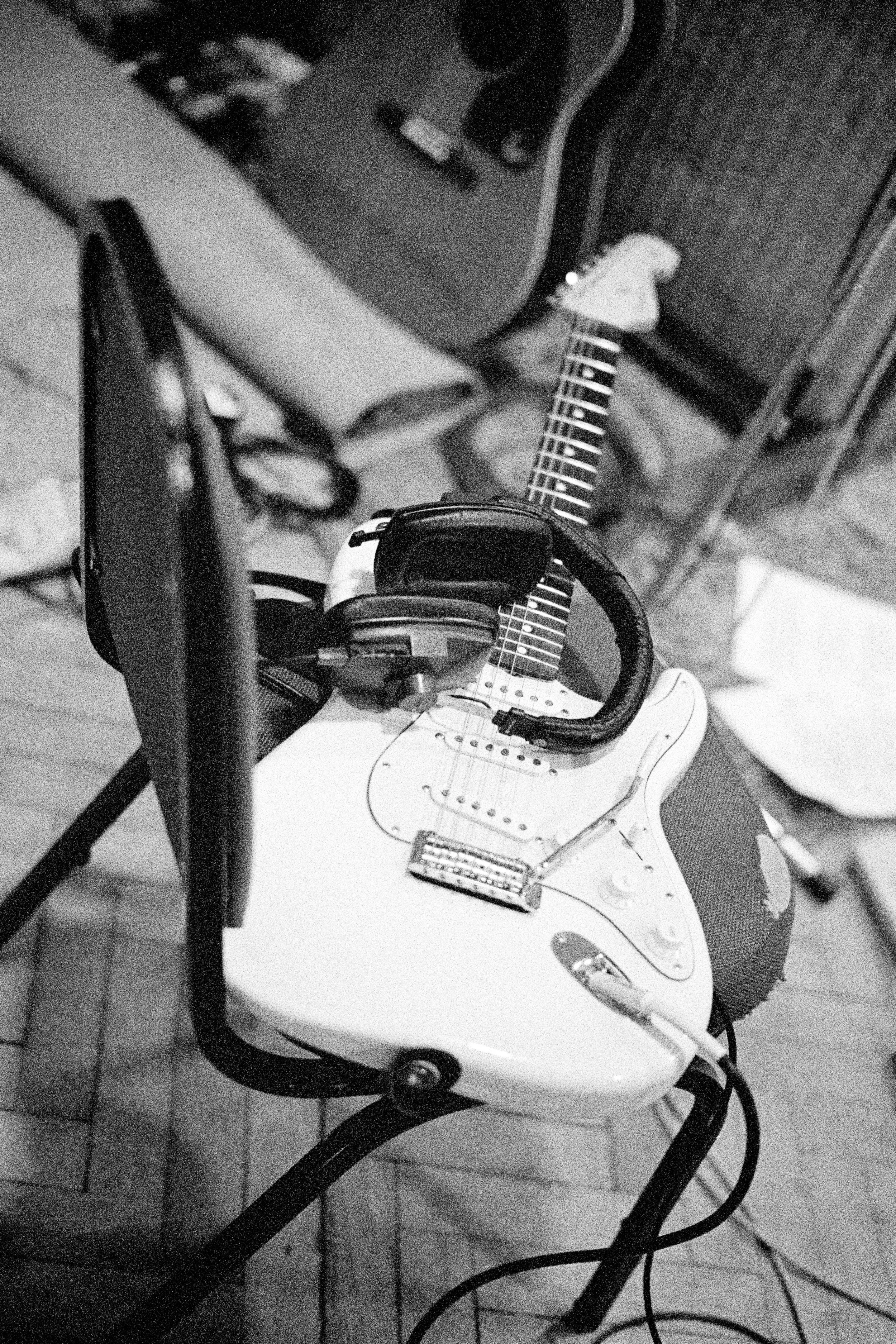 Black and white photo of an electric guitar on a chair, with headphones draped over it, and a phone mounted on a stand nearby.