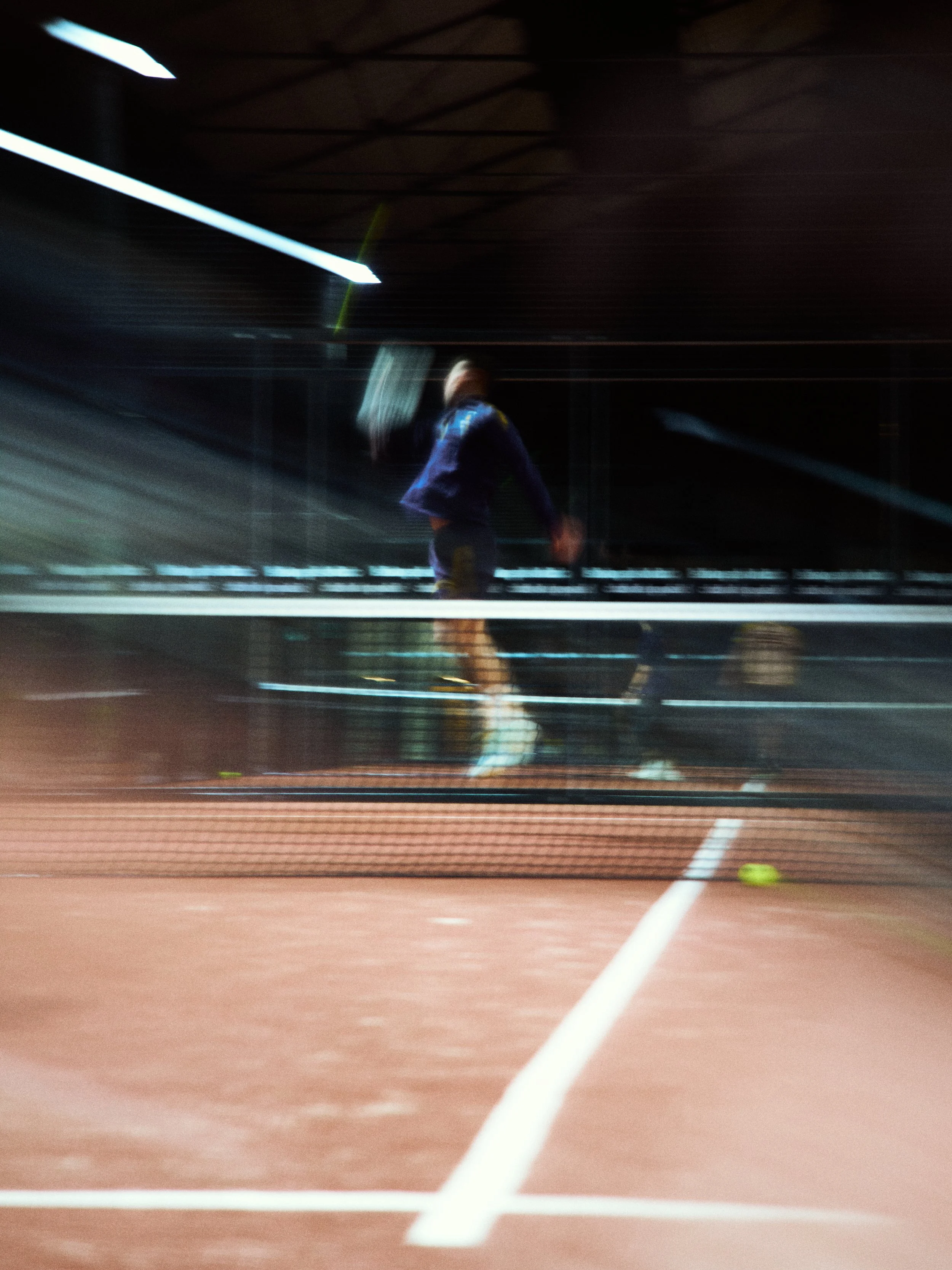 A person playing tennis on an indoor court at night, with motion blur showing movement.