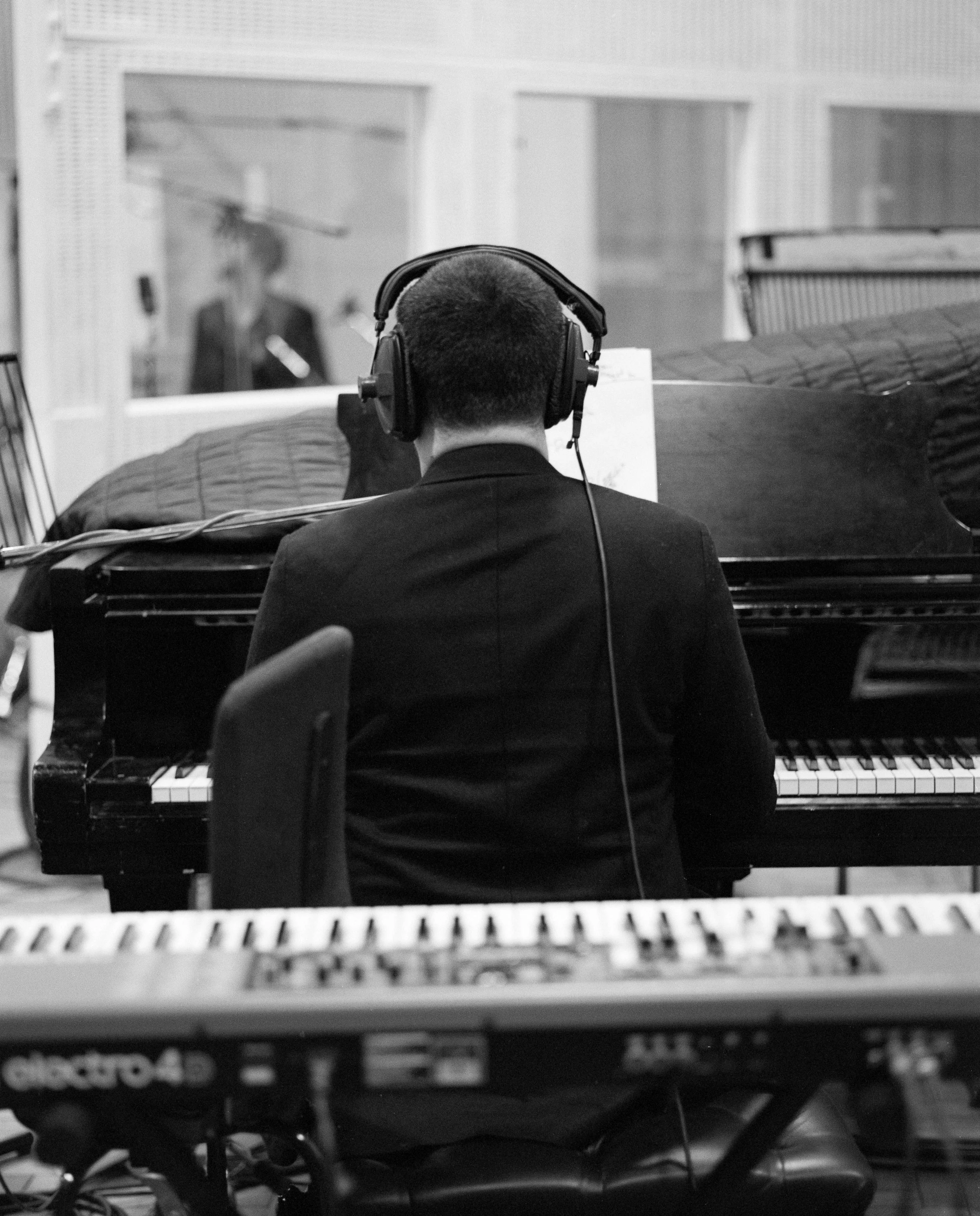 A person wearing headphones playing a piano in a recording studio, viewed from behind, with a reflection of another person in the glass window.