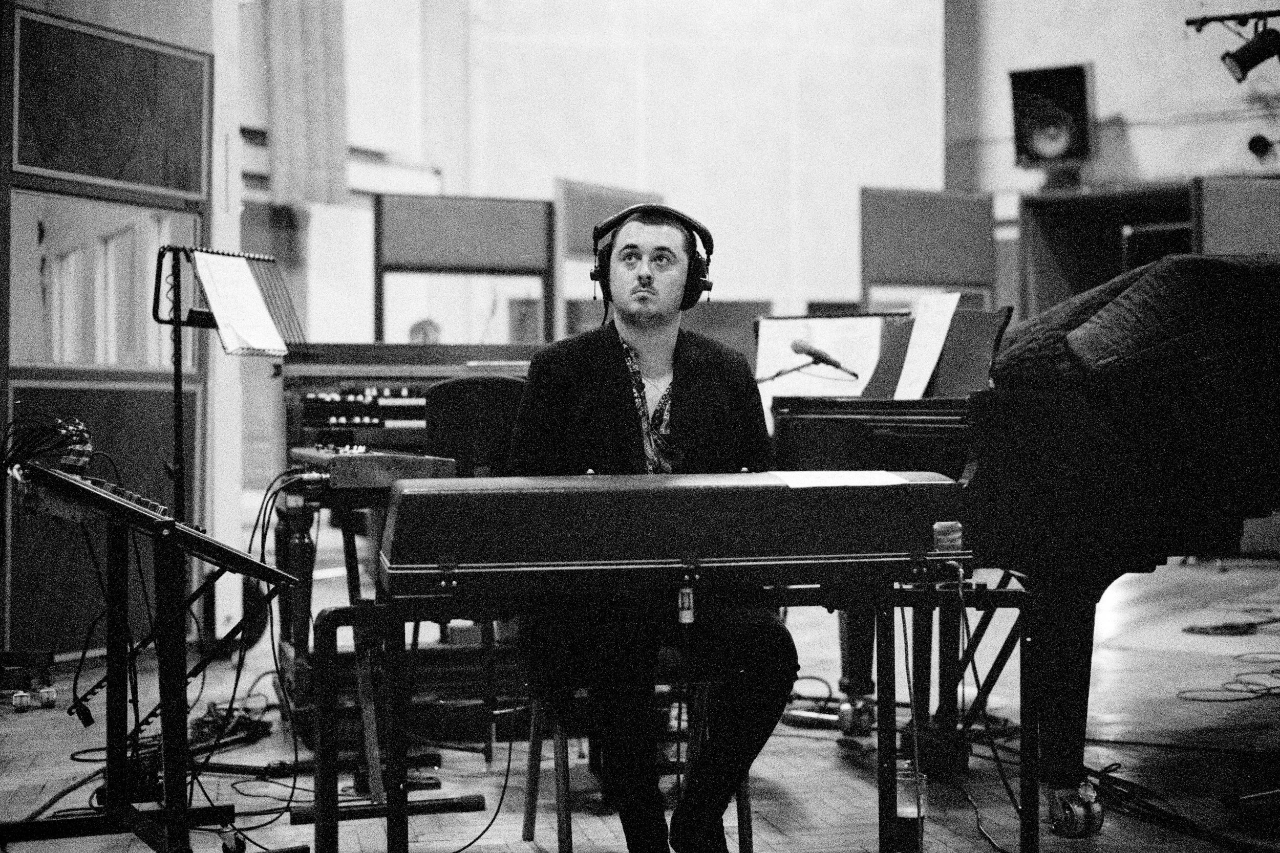A man in a recording studio sitting at a keyboard, wearing headphones, with music equipment and music sheets around him.