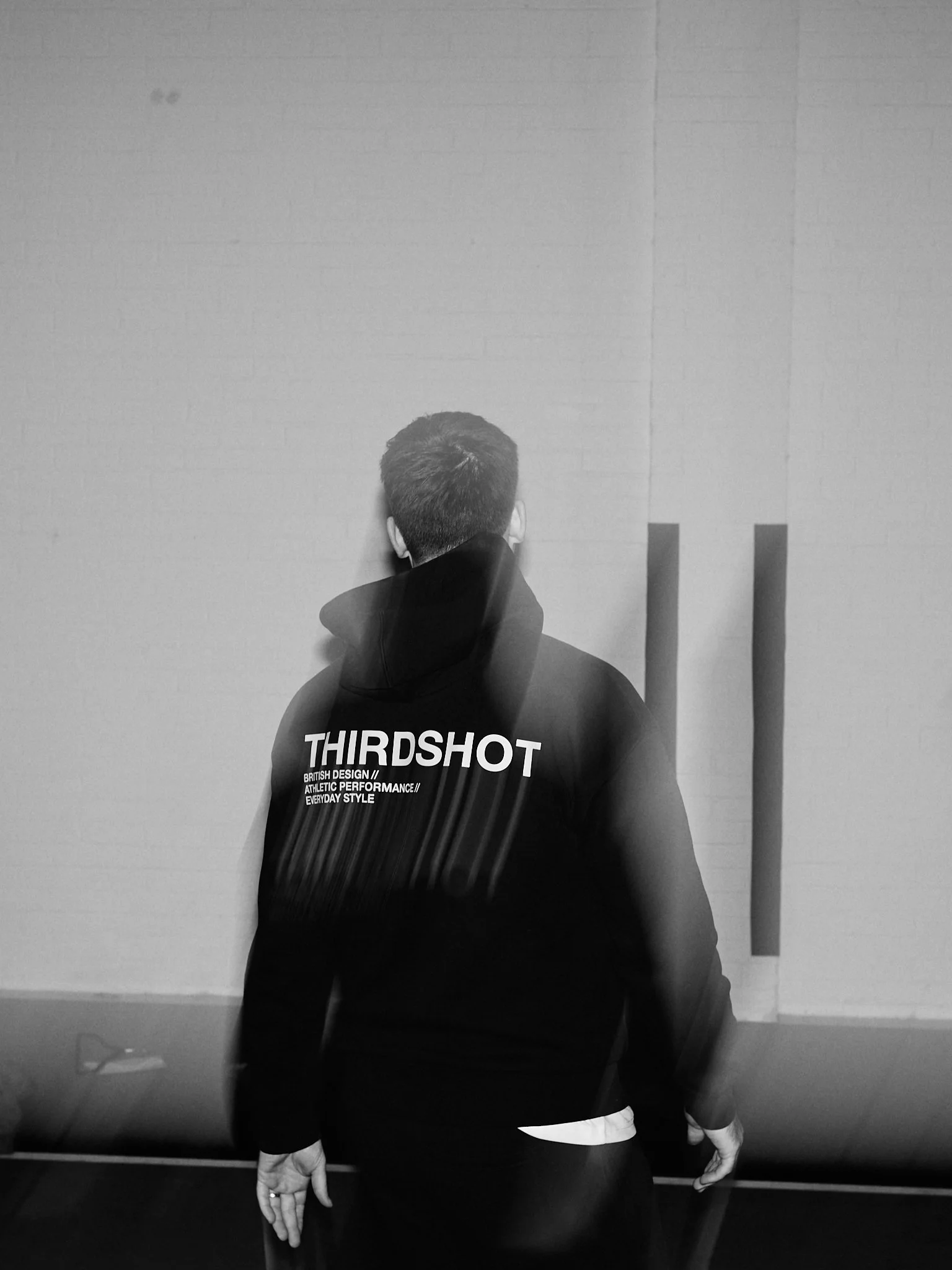A person with short hair, wearing a black hoodie with the word "THIRDSHOT" on the back, stands with their back to the camera, facing a plain brick wall.