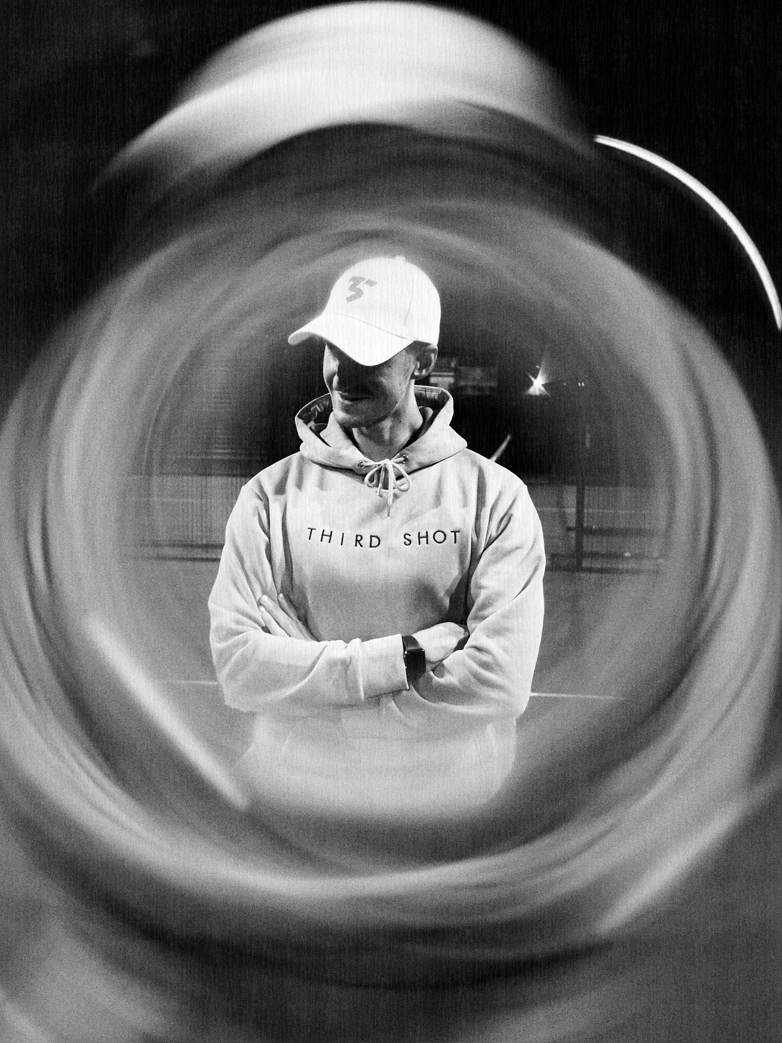 A person wearing a cap and hoodie stands with arms crossed, seen through a circular object, at night.