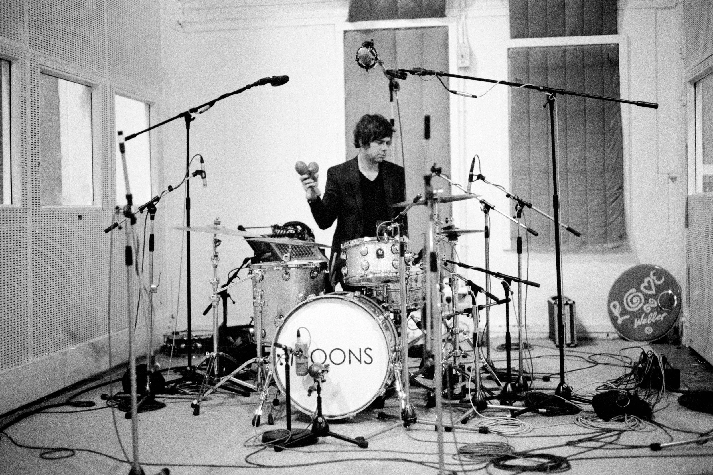 A musician is standing behind a drum set in a recording studio, surrounded by microphones and cables, holding a maraca in one hand and a tablet in the other.