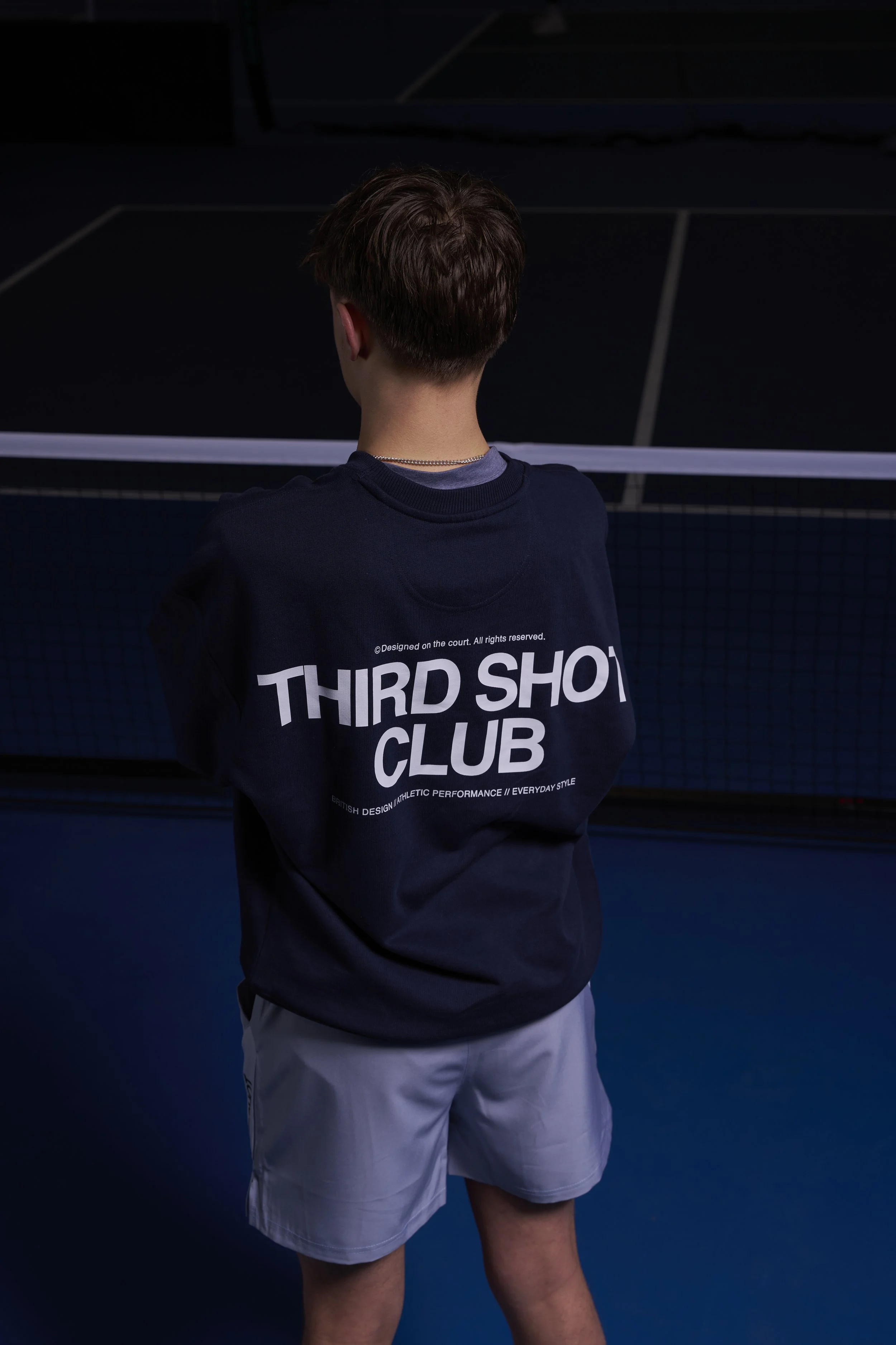 A person with short brown hair stands on an indoor tennis court, facing away from the camera, wearing a navy blue sweatshirt with 'Third Shot Club' written on the back, and light gray shorts.