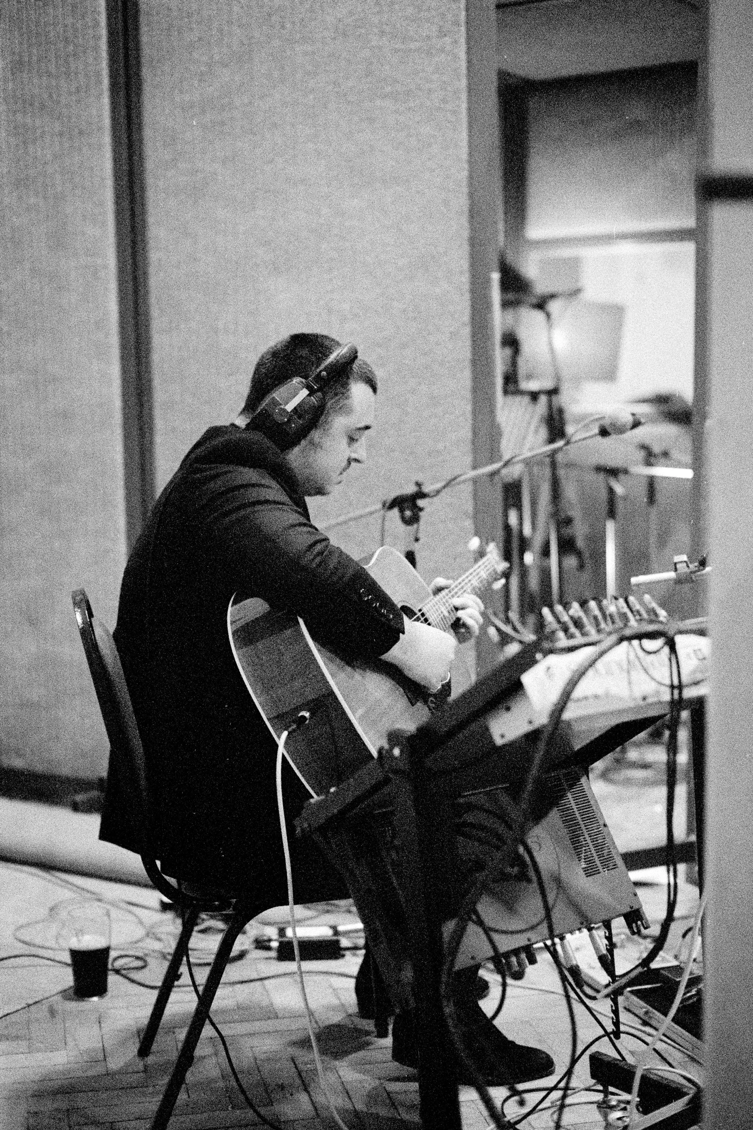 A man wearing headphones is sitting on a chair playing an acoustic guitar in a recording studio, with sound equipment and microphones around him.