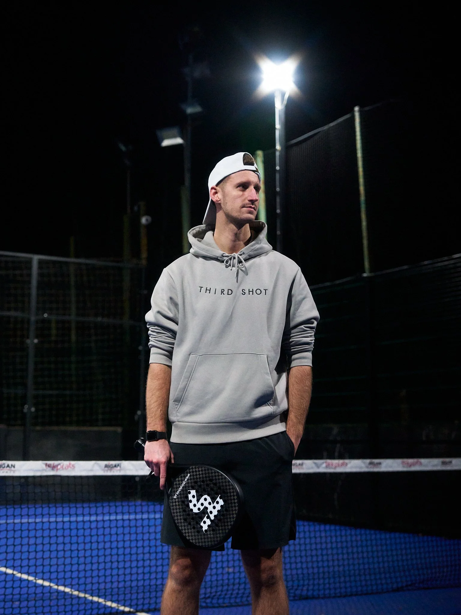 A young man standing on a nighttime padel tennis court under bright floodlights, dressed in casual athletic clothing, holding a padel racket.