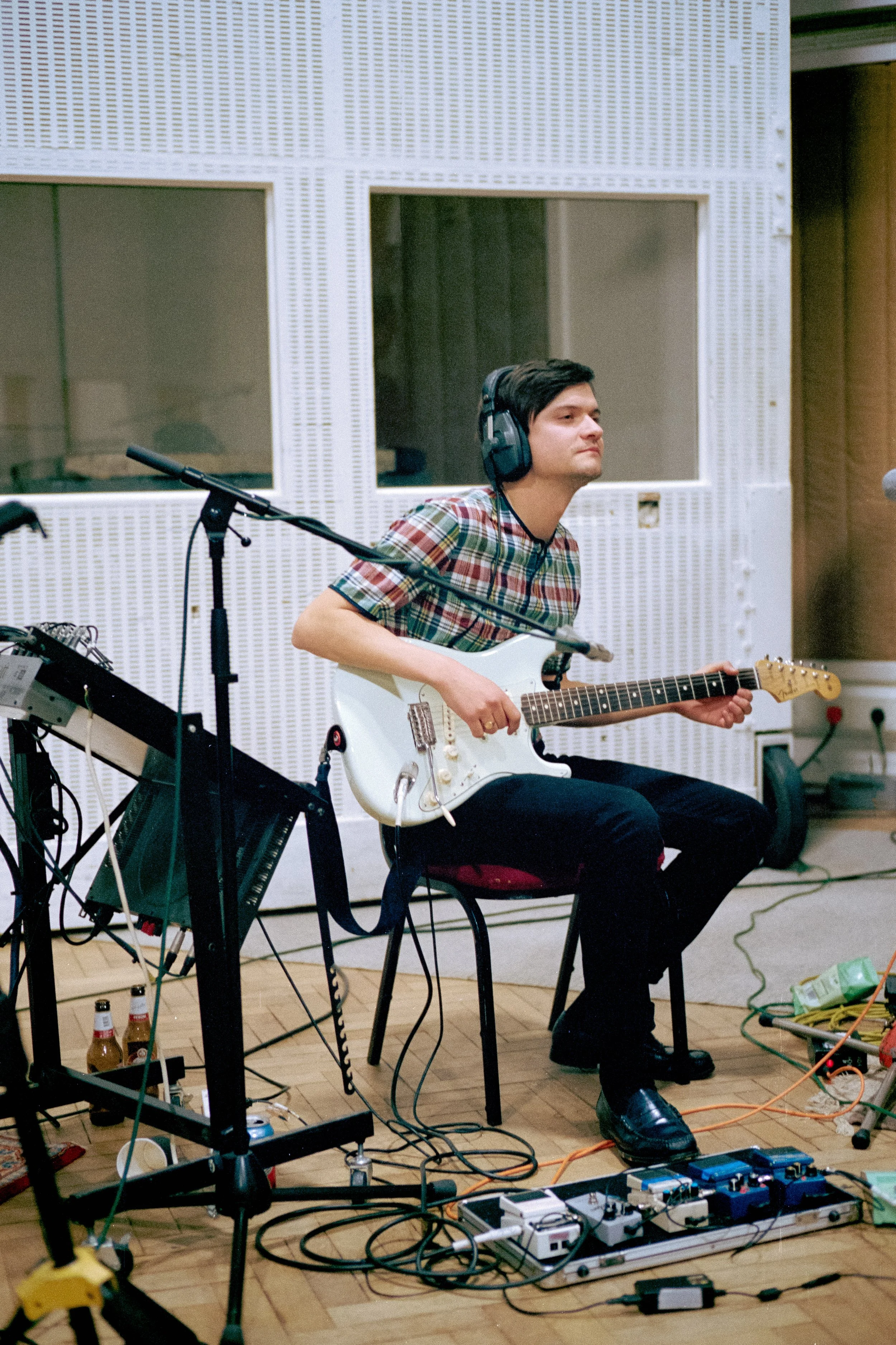 A man in a plaid shirt playing an electric guitar in a recording studio, wearing large headphones.