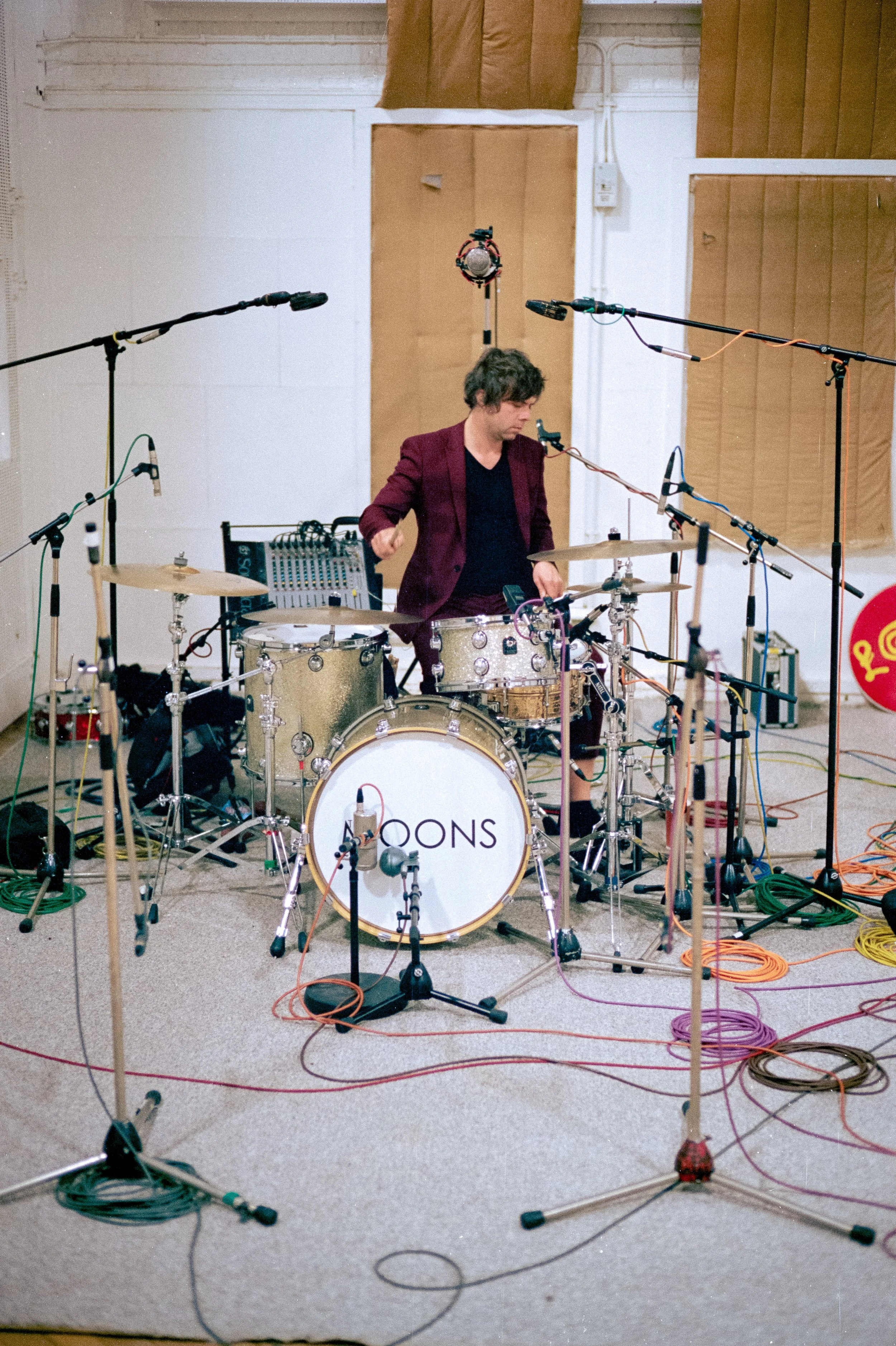 A musician playing drums in a recording studio with multiple microphones and equipment around.