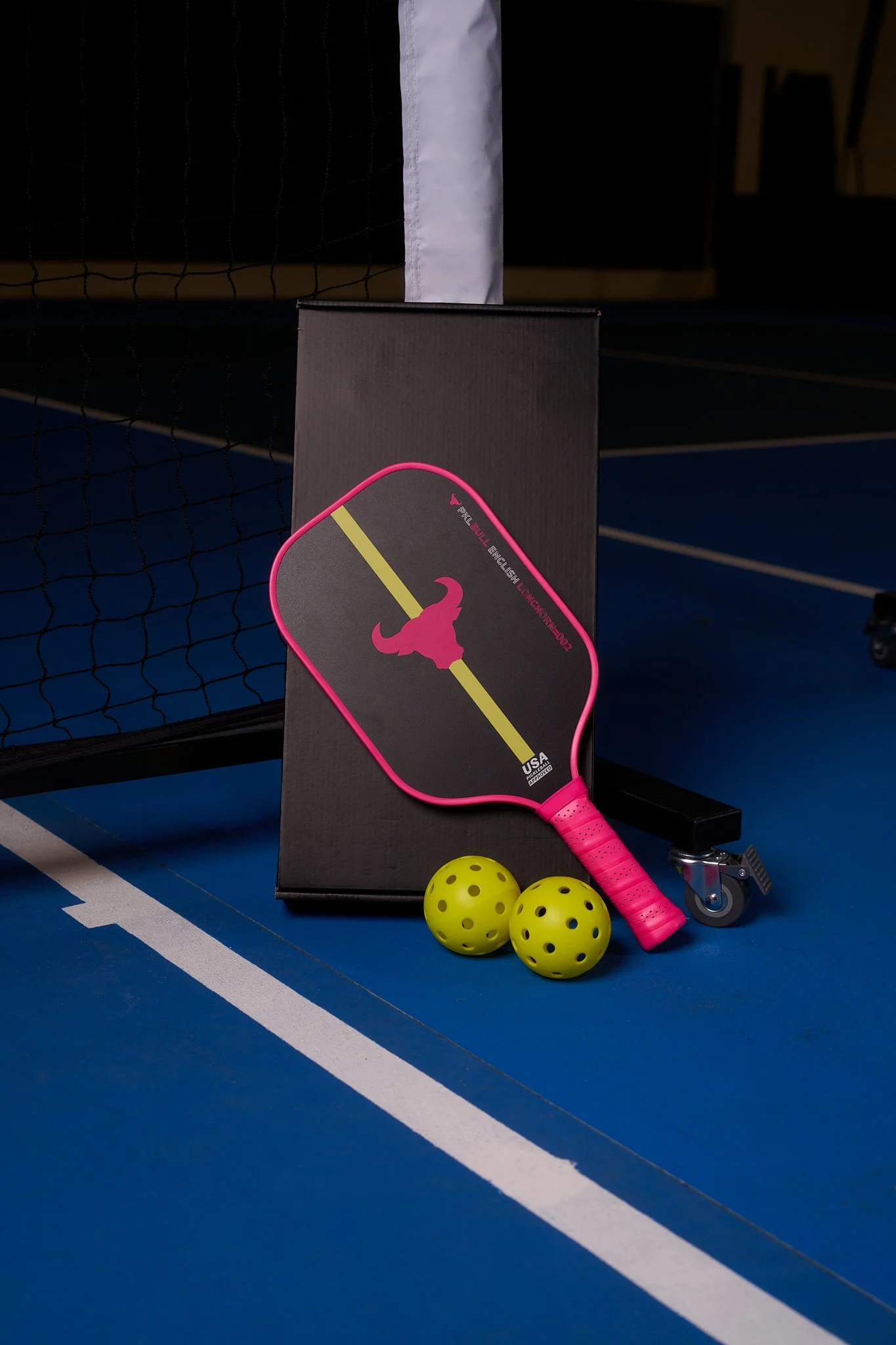 A pink pickleball paddle with a black face and yellow design along with two yellow pickleballs with black holes, resting on a blue indoor pickleball court near a net and black box.