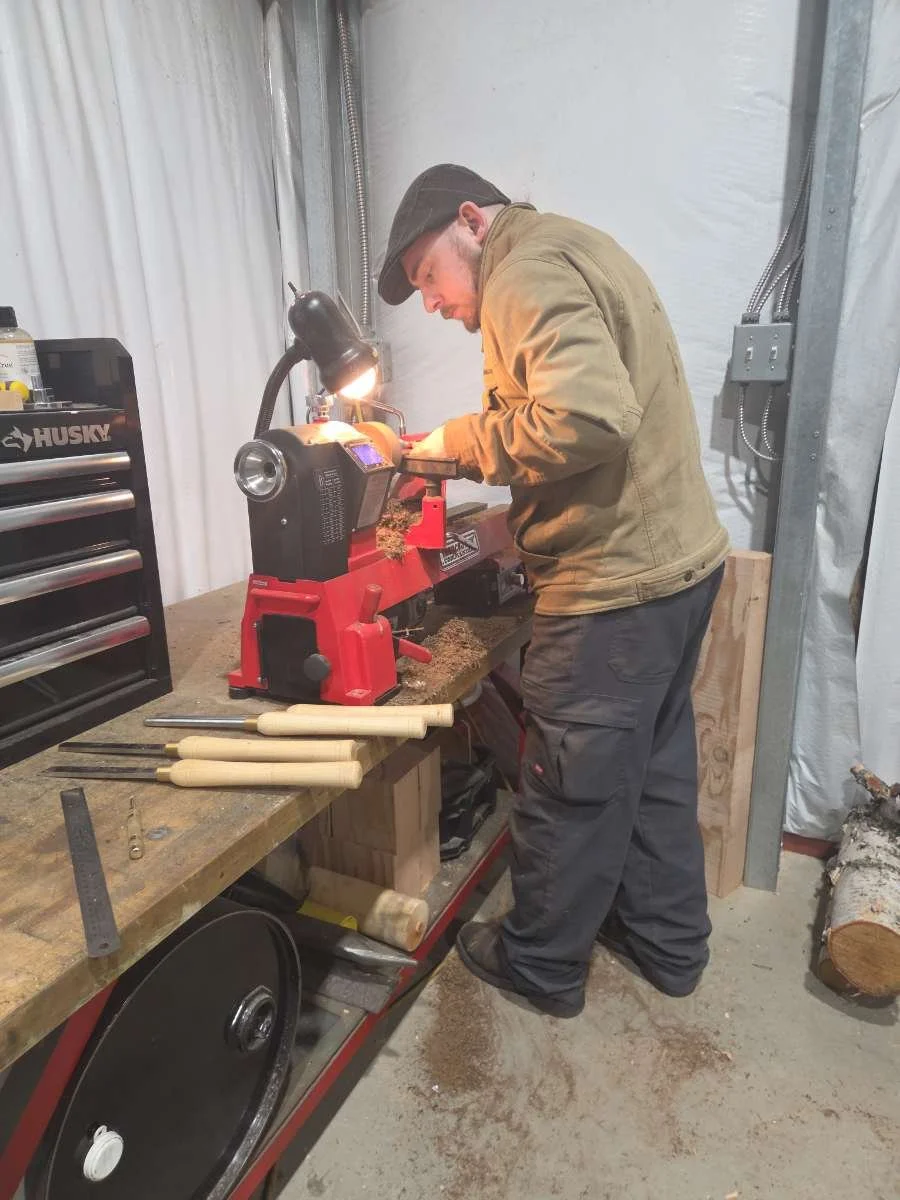 Man working on a wood lathe in a workshop, wearing a cap and beige jacket, with wooden tools and logs nearby.
