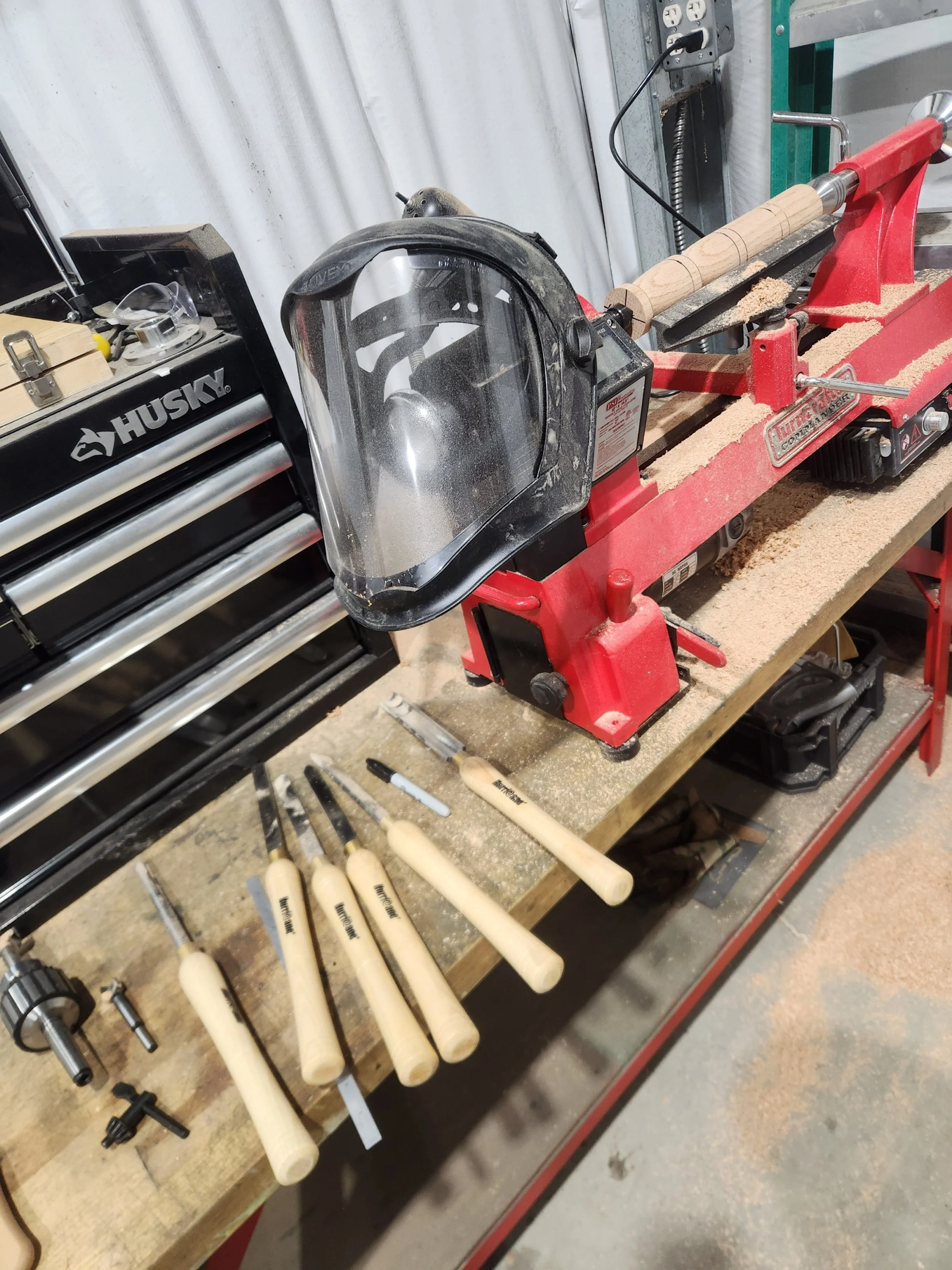 Workbenches with woodworking tools, a red mini lathe with a partially turned wooden piece, and a set of carving chisels with light-colored handles in a woodworking workshop.