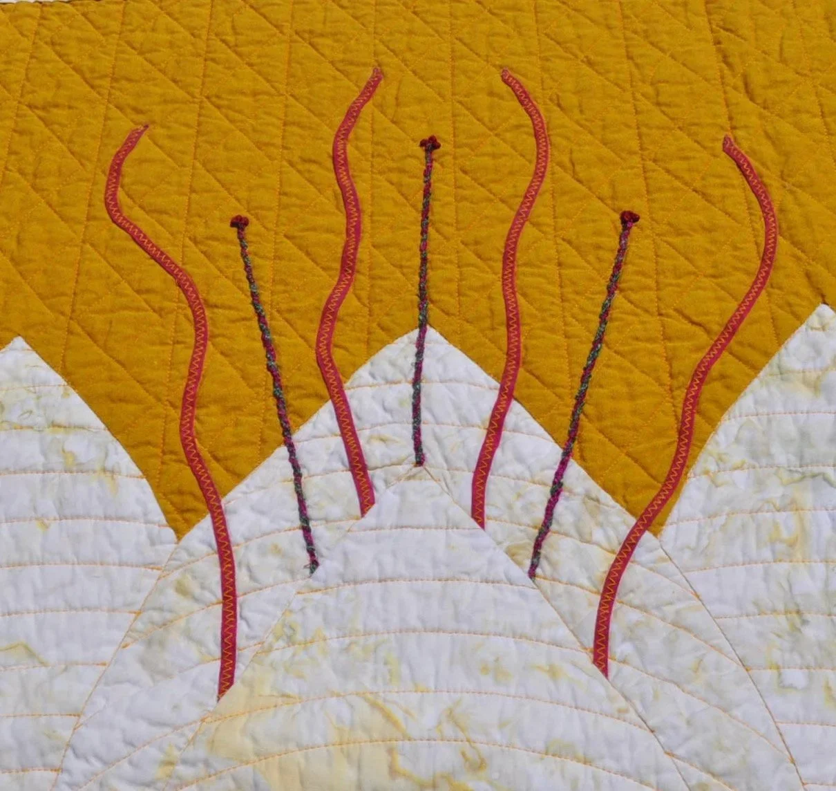 Close-up view of a quilted fabric with embroidered design resembling mountain peaks and red wavy lines representing streams or smoke.