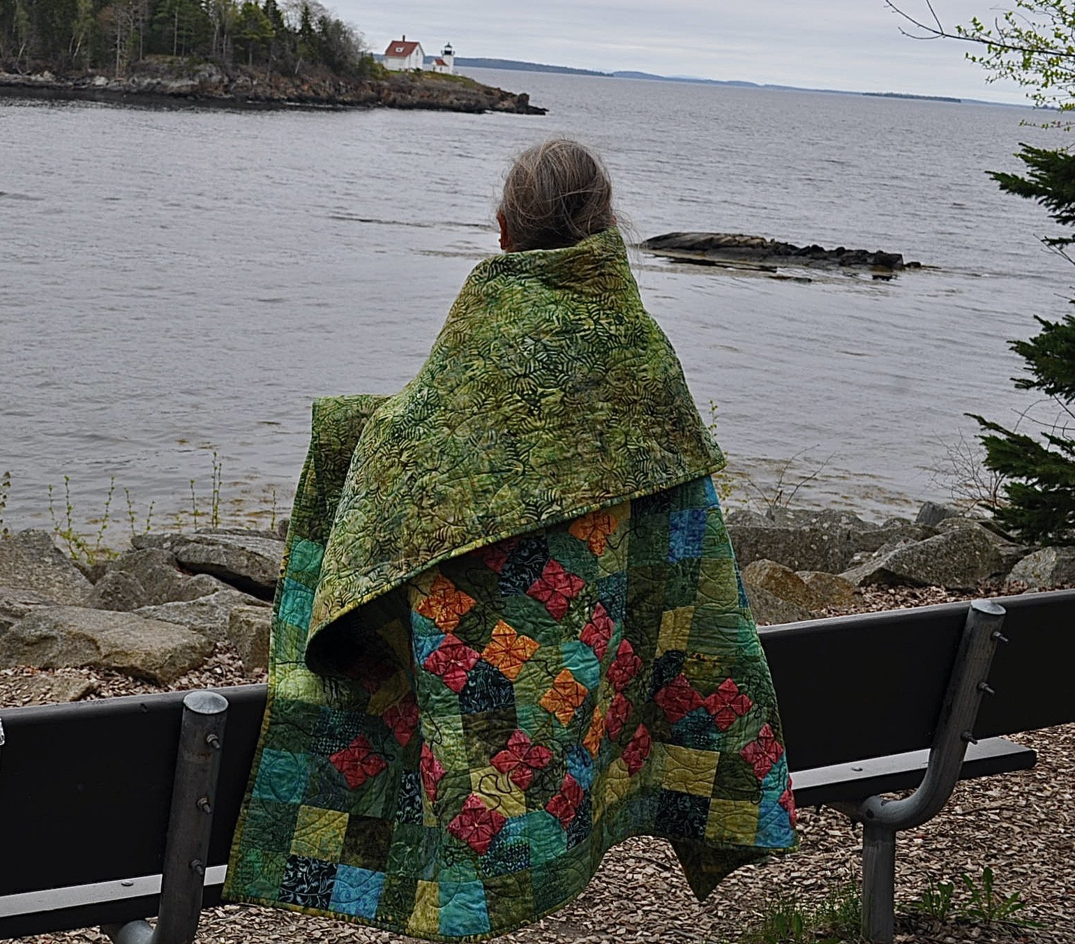 A woman sitting on a park bench wrapped in a colorful quilt, overlooking a body of water with a rocky shore and a lighthouse in the distance.