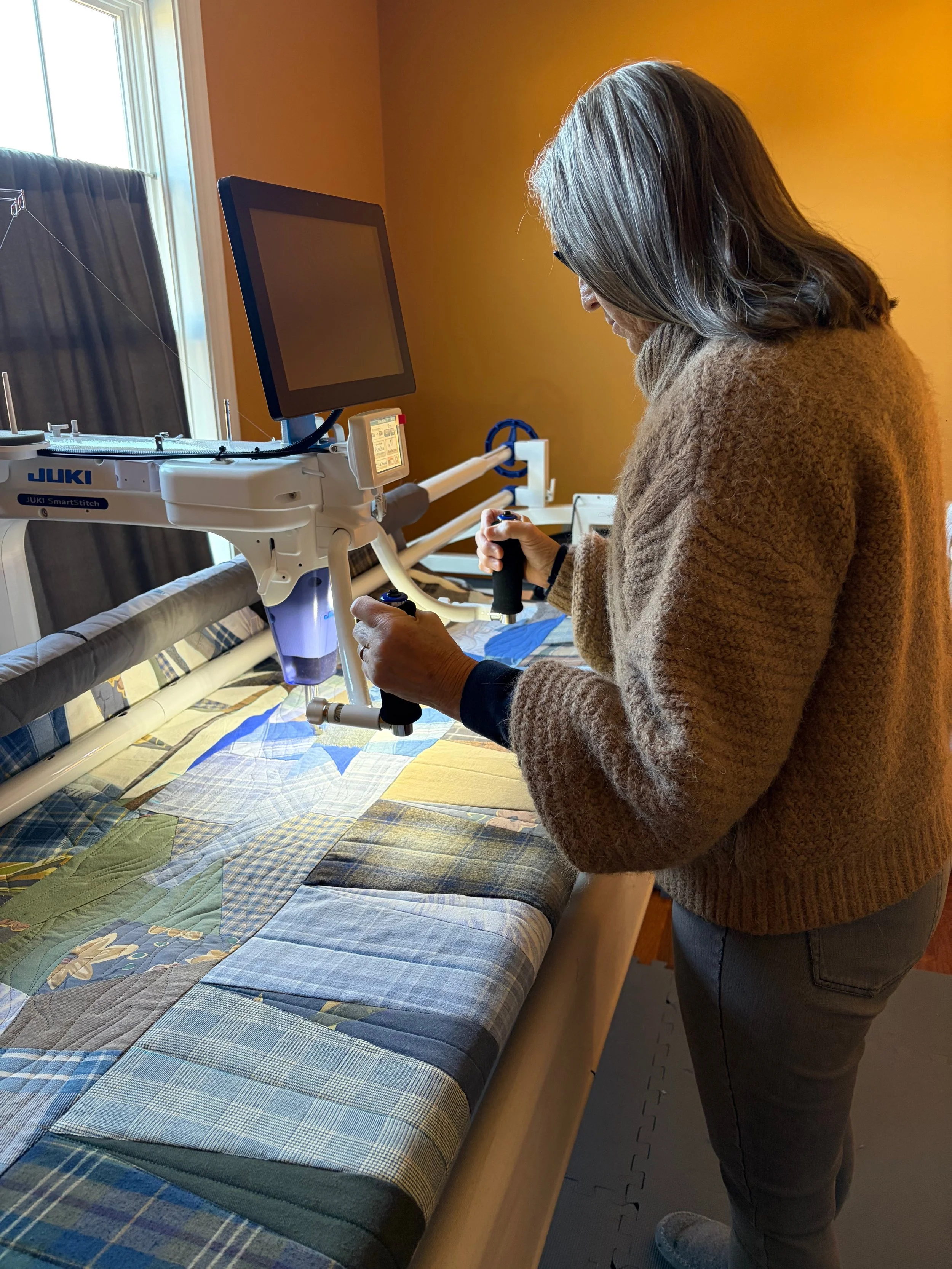 A woman in a brown sweater working with a Juki sewing machine and a computer on a sewing table, with a quilted fabric underneath.