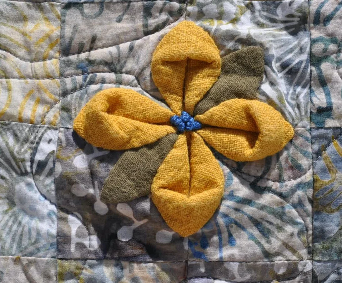 A fabric flower with yellow and gray fabric petals sewn onto a quilted surface.