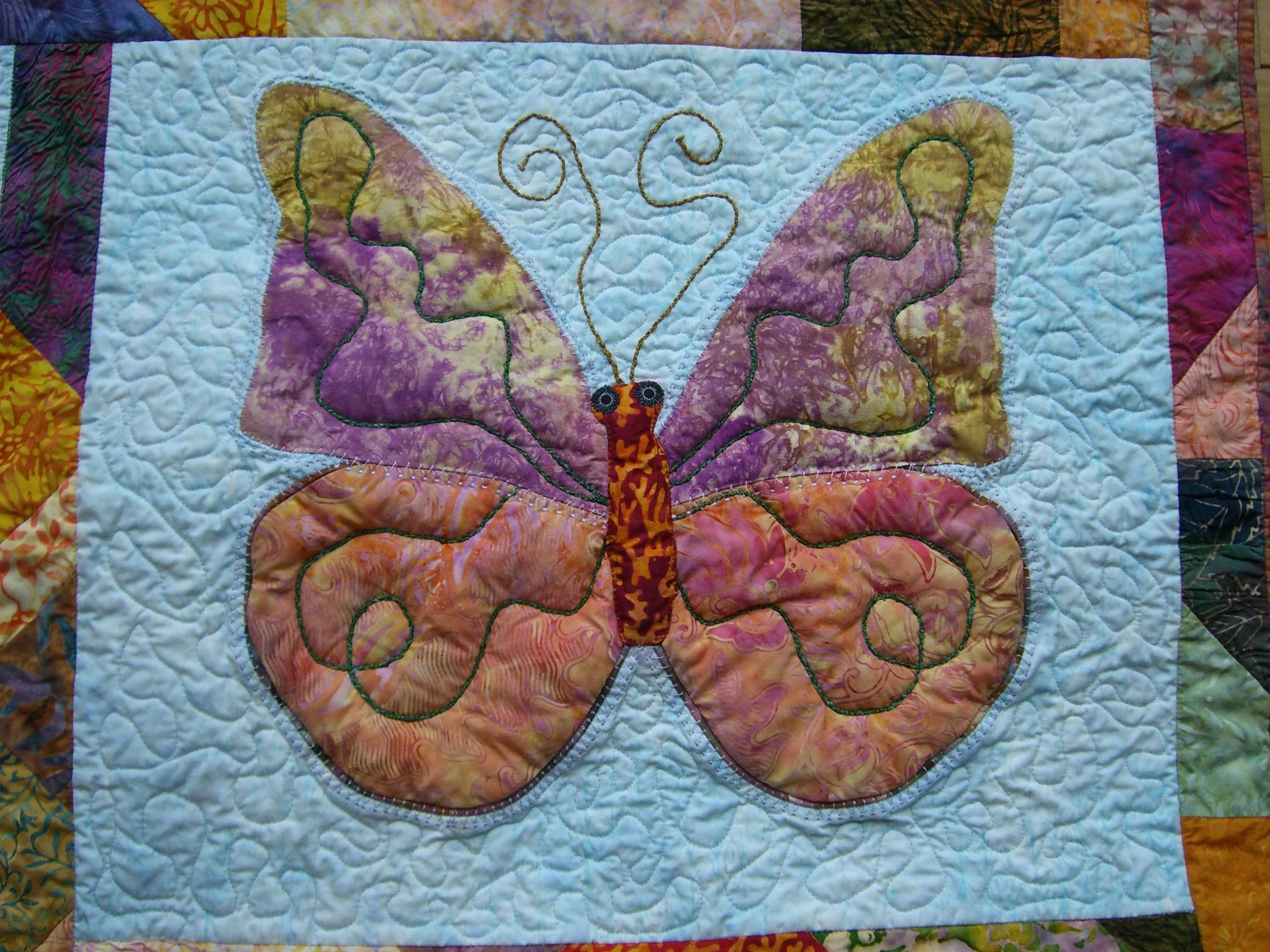 A quilt with a butterfly design, featuring colorful fabric wings and embroidered details, on a multicolored patchwork background.
