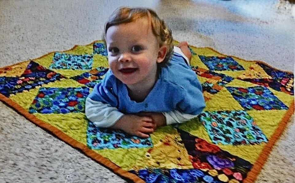 A young child with curly red hair lying on a colorful quilt on a carpeted floor, smiling at the camera.