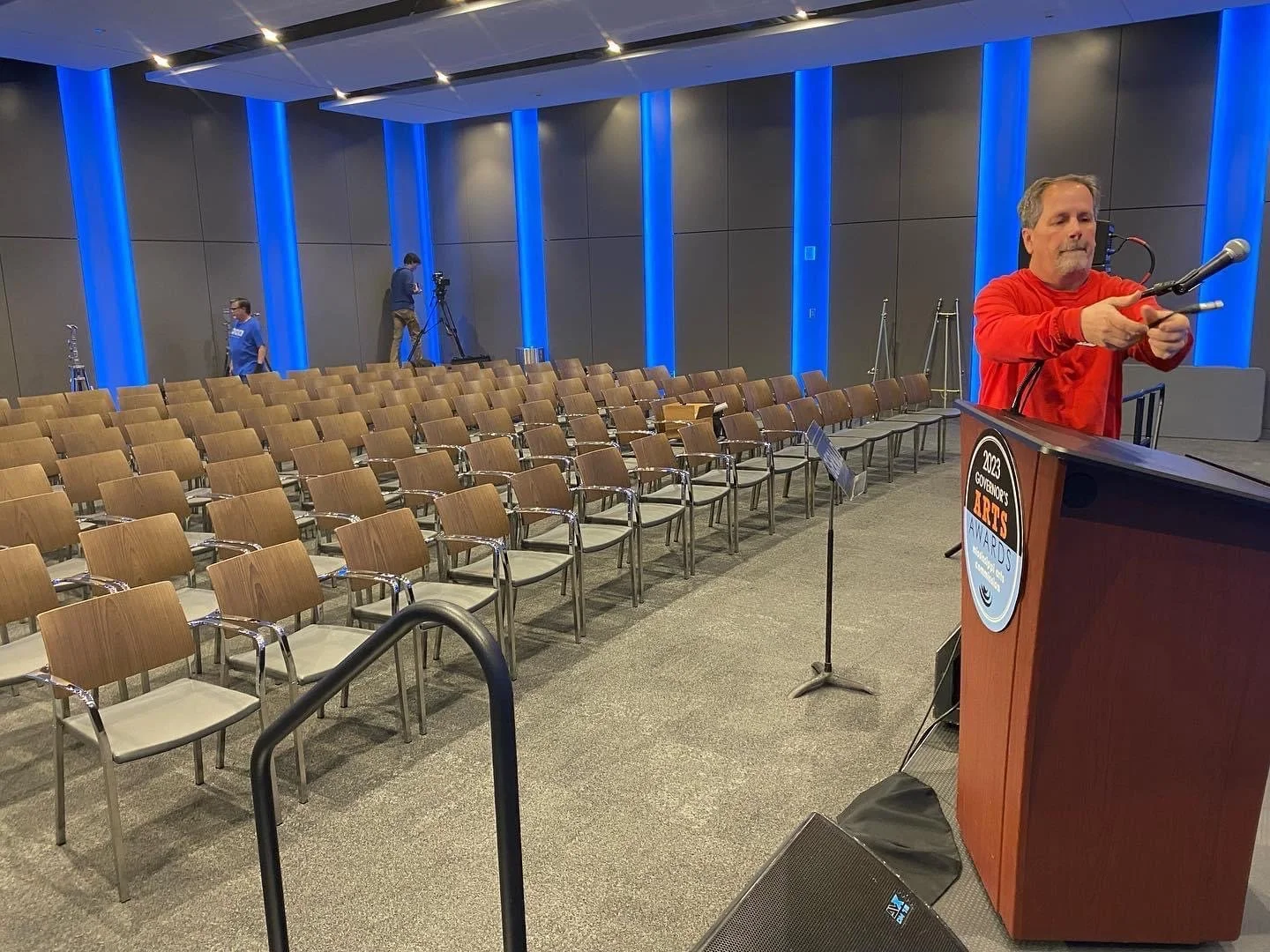 An empty auditorium with rows of brown chairs, a man in a red shirt setting up audio equipment at a podium, and two technicians with cameras in the background.