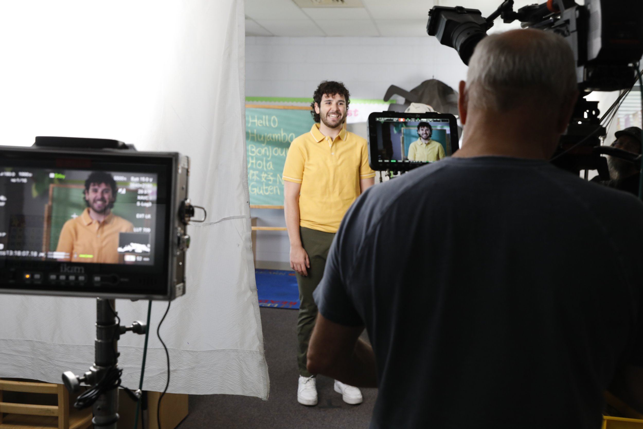 A man in a yellow shirt standing in front of a green chalkboard with greeting words in multiple languages is being filmed by a camera crew in a studio setting.