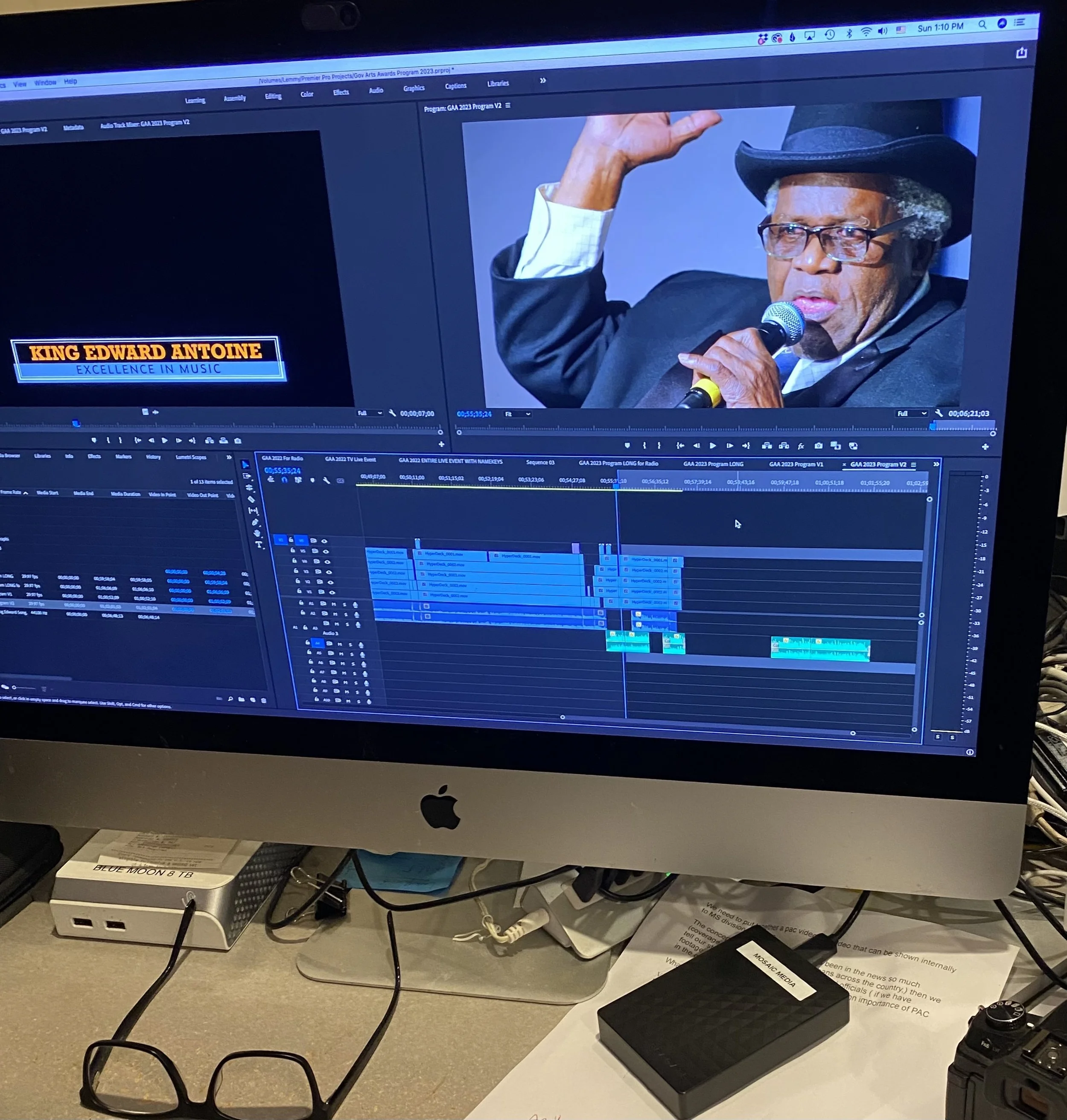 Computer monitor displaying a video editing software with a timeline and a photo of a man speaking into a microphone, wearing glasses, a black hat, and a suit. Text on the video says 'KING EDWARD ANTOINE EXCELLENCE IN MUSIC'.
