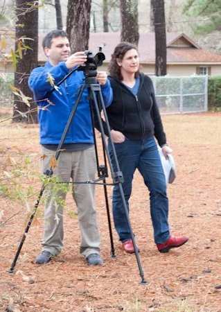 Two people standing outdoors on a dirt surface, with the man operating a camera on a tripod and the woman holding papers, in a wooded area with a house and a fence in the background.