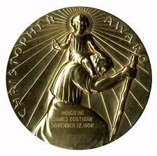 Gold-colored medallion featuring a figure kneeling in prayer, with rays emanating from behind and Latin inscriptions at the top, honoring Padre Pio.