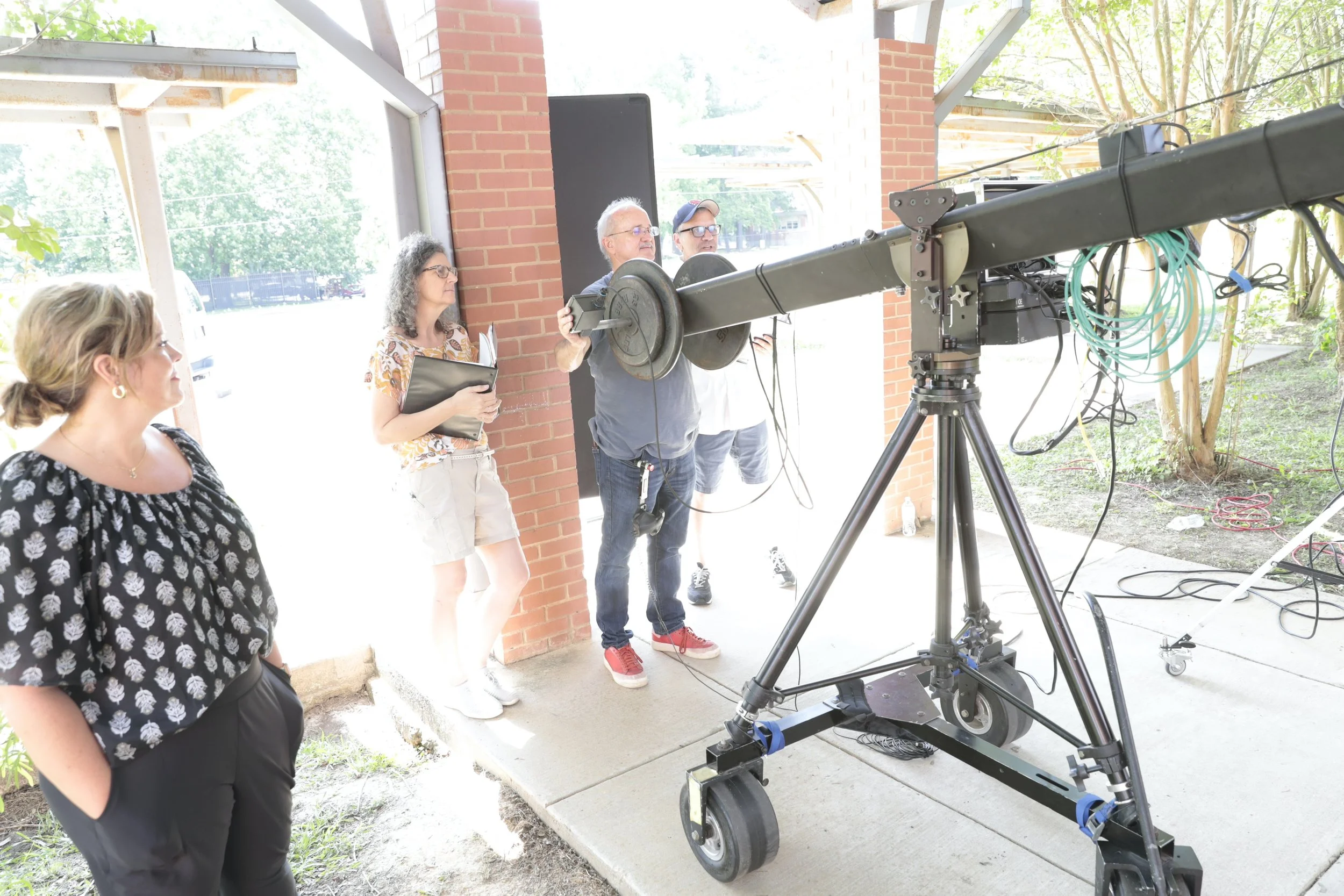 Filmmaking crew setting up a large camera on a dolly outdoors during the day, with some people standing nearby near a brick wall.