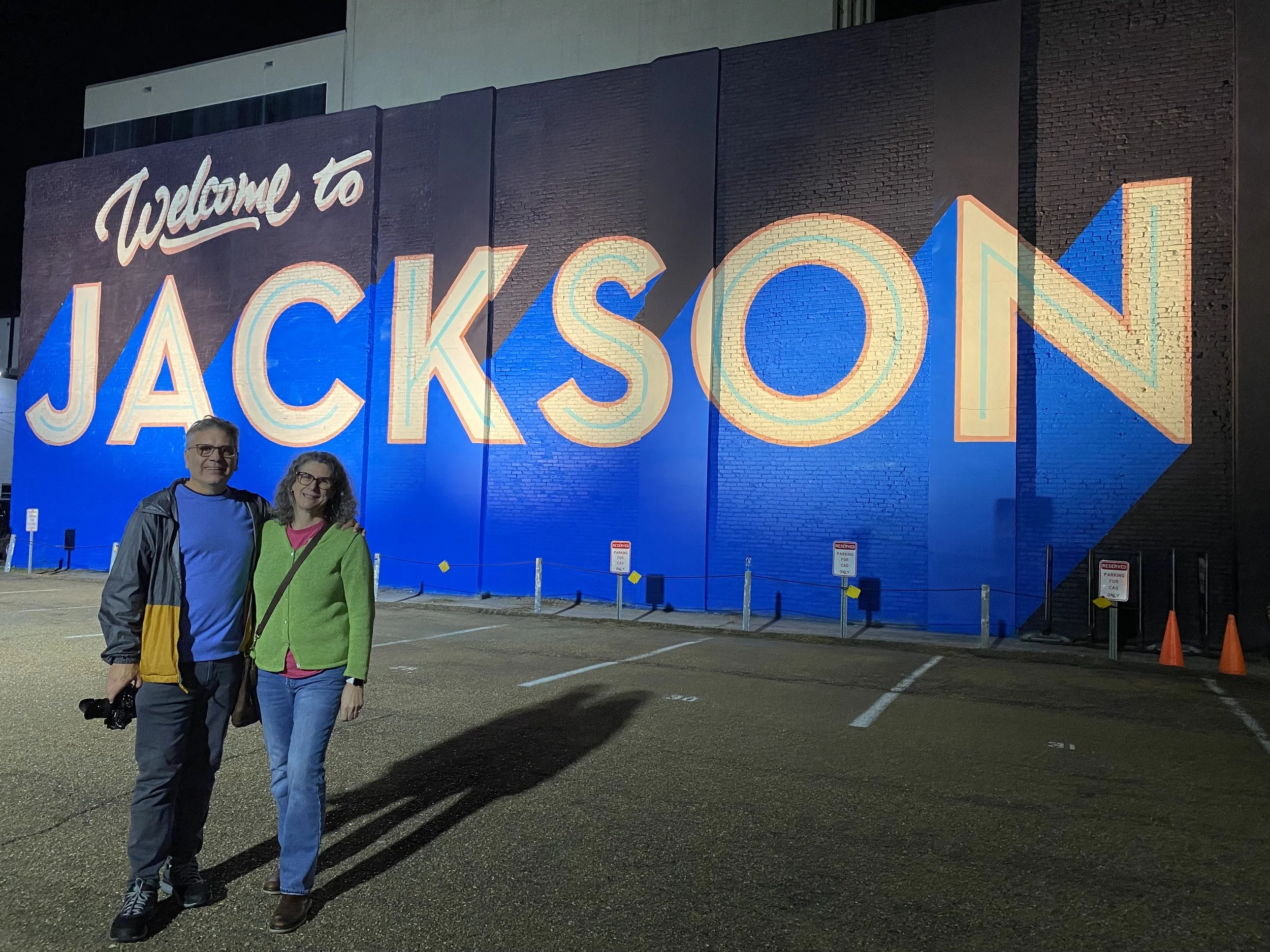 A man and woman standing together in a parking lot at night, smiling for the photo. Behind them is a large mural on a brick wall that says 'Welcome to JACKSON' in colorful letters, illuminated by lights.