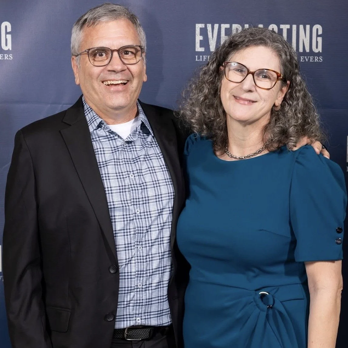 A man and woman standing together in front of a backdrop with the word "EVERLASTING" on it, smiling and looking at the camera.