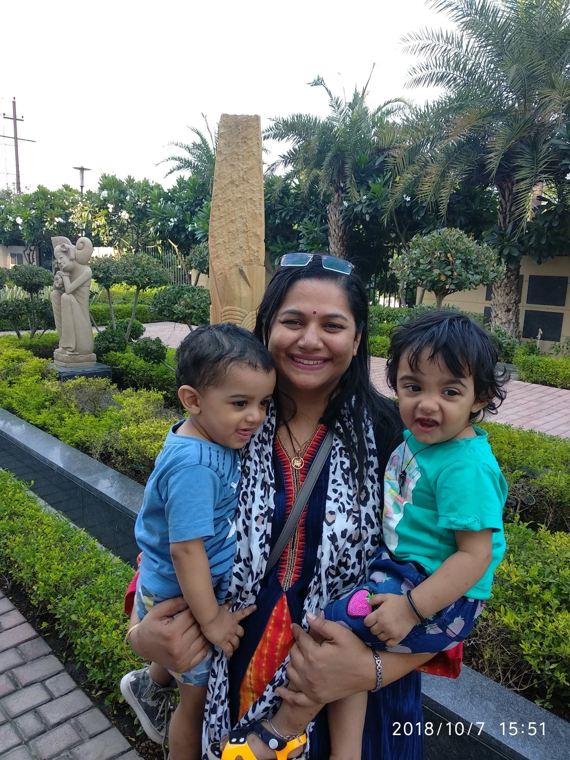 A woman holding two young children outdoors in a garden with sculptures and palm trees. All are smiling.