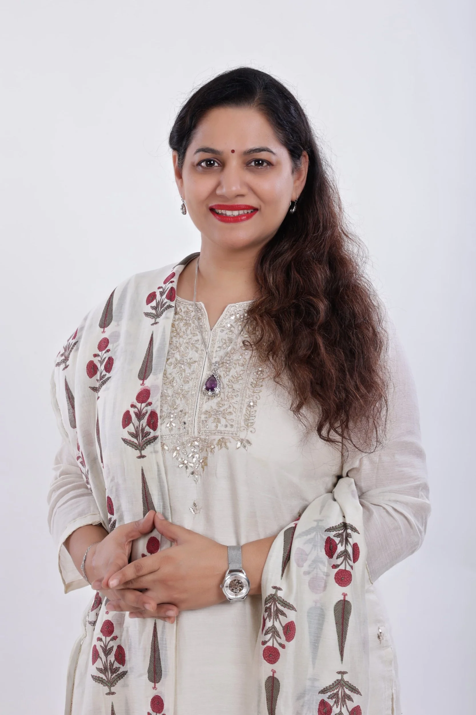 A woman with long, wavy brown hair smiling, wearing traditional Indian attire with embroidery and a matching scarf, jewelry, and a wristwatch, standing against a plain white background.
