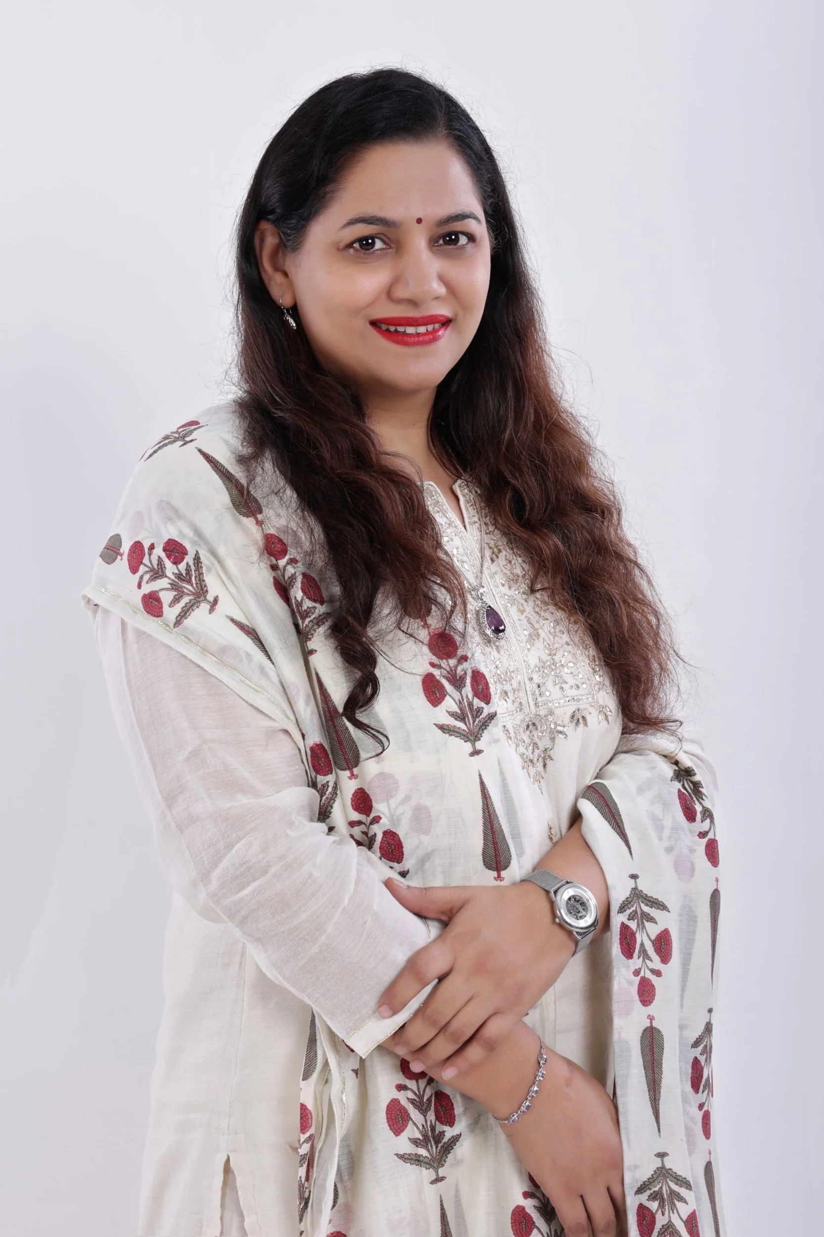 A woman with long, wavy dark hair and a bindi on her forehead poses with a smile, wearing a cream-colored embroidered kurta with red and brown floral designs, accessorized with a wristwatch and jewelry, against a plain white background.