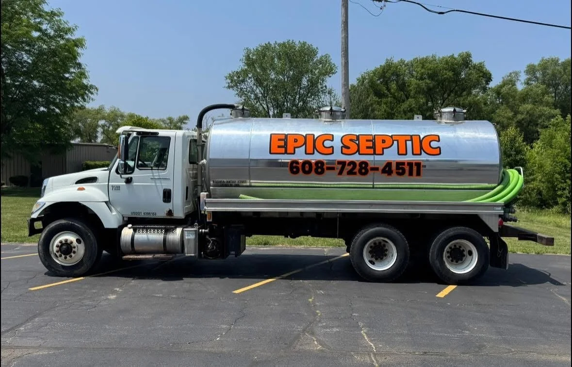 A septic service truck parked in a parking lot with trees and a blue sky in the background. The truck has a large silver tank with orange and black lettering that reads "EPIC SEPTIC" and a phone number, along with green hoses attached.
