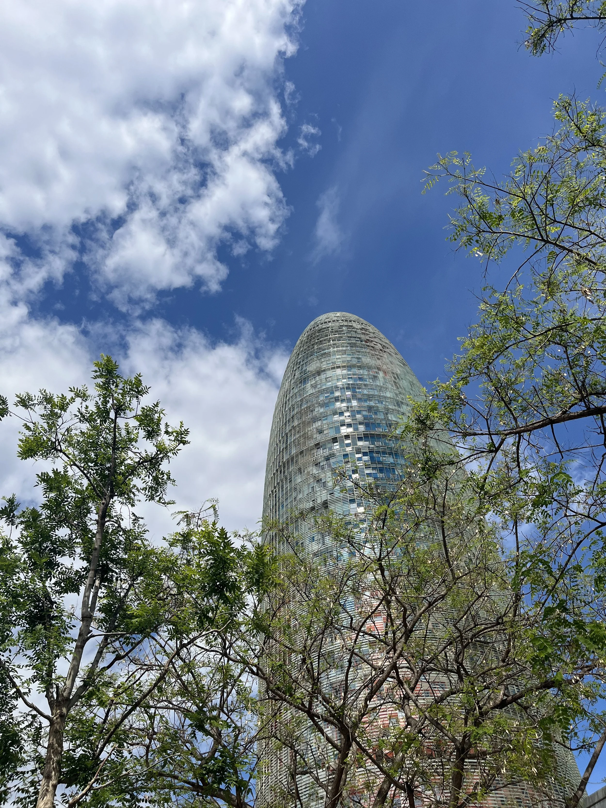 Looking up at the Torre Glòries building in Barcelona, Spain, with trees in the foreground and a partly cloudy sky overhead.