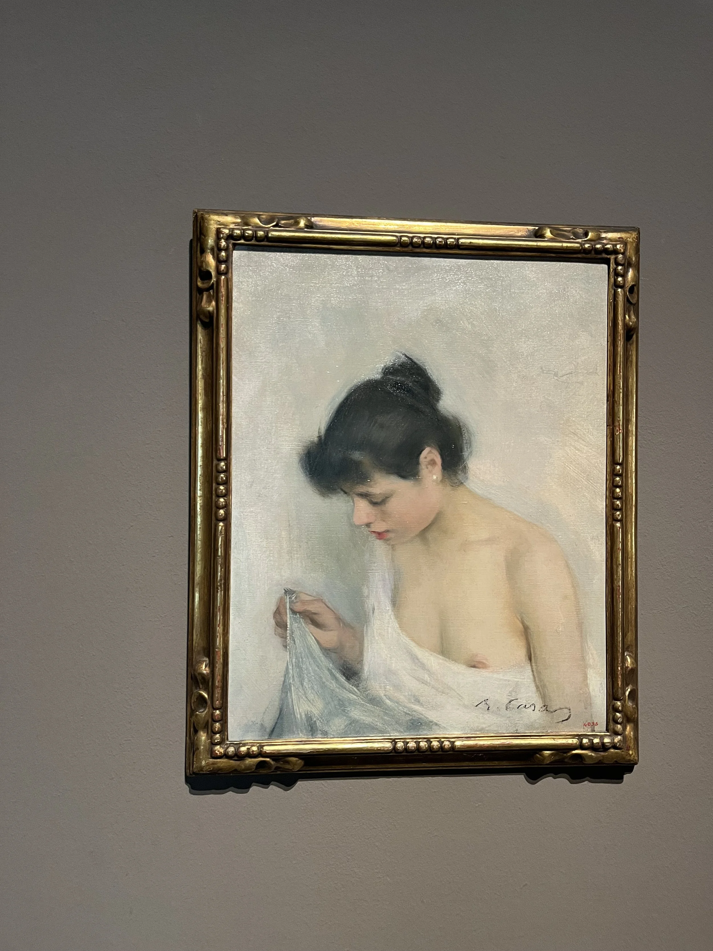A realistic painting of a young woman with dark hair tied in a bun, looking down while holding a light-colored cloth. The woman appears to be nude from the waist up, with a soft and delicate expression. The painting is framed in an ornate gold frame.