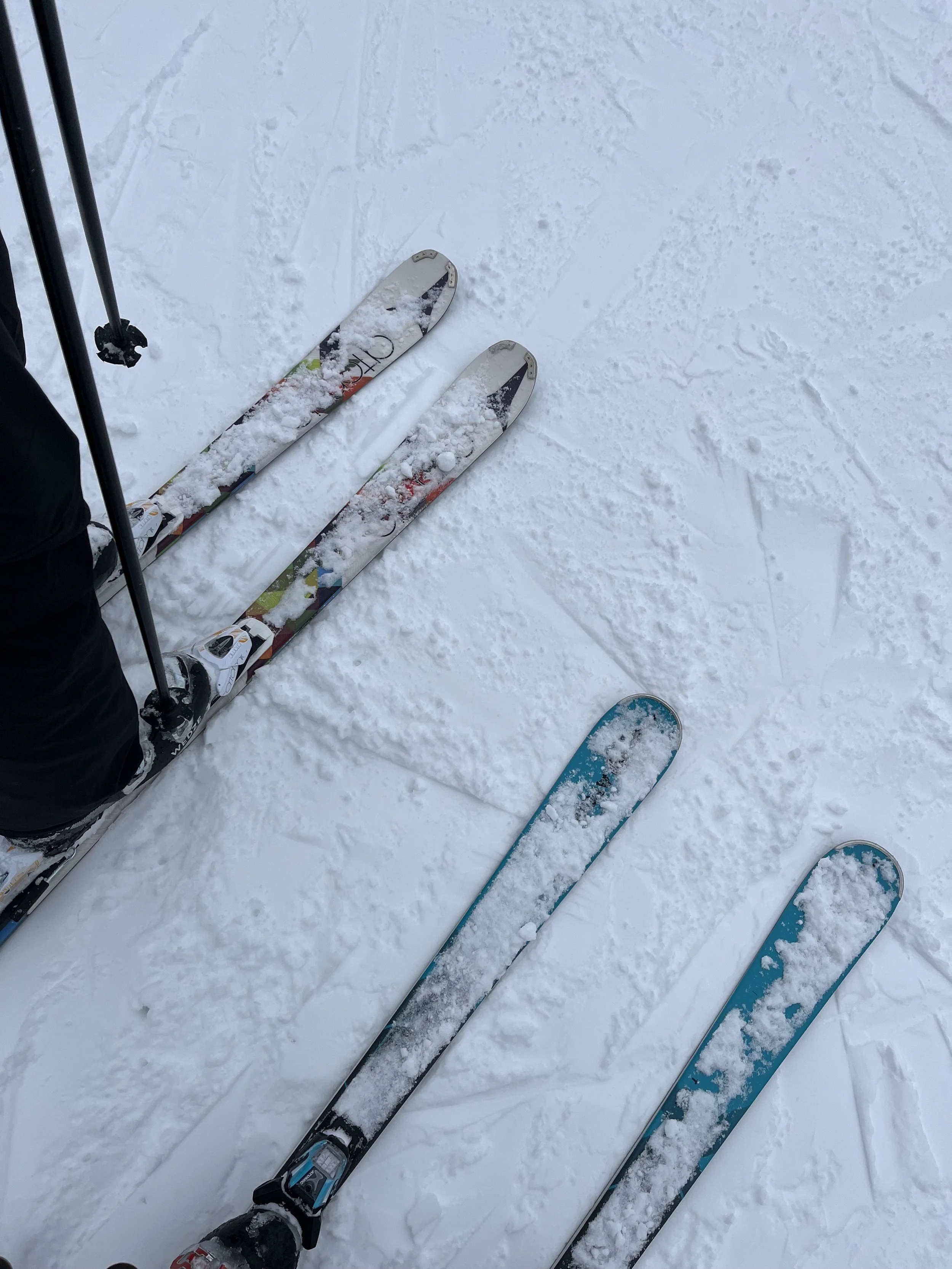 Two pairs of skis resting on snow, with the person's legs and ski poles visible on the left side.