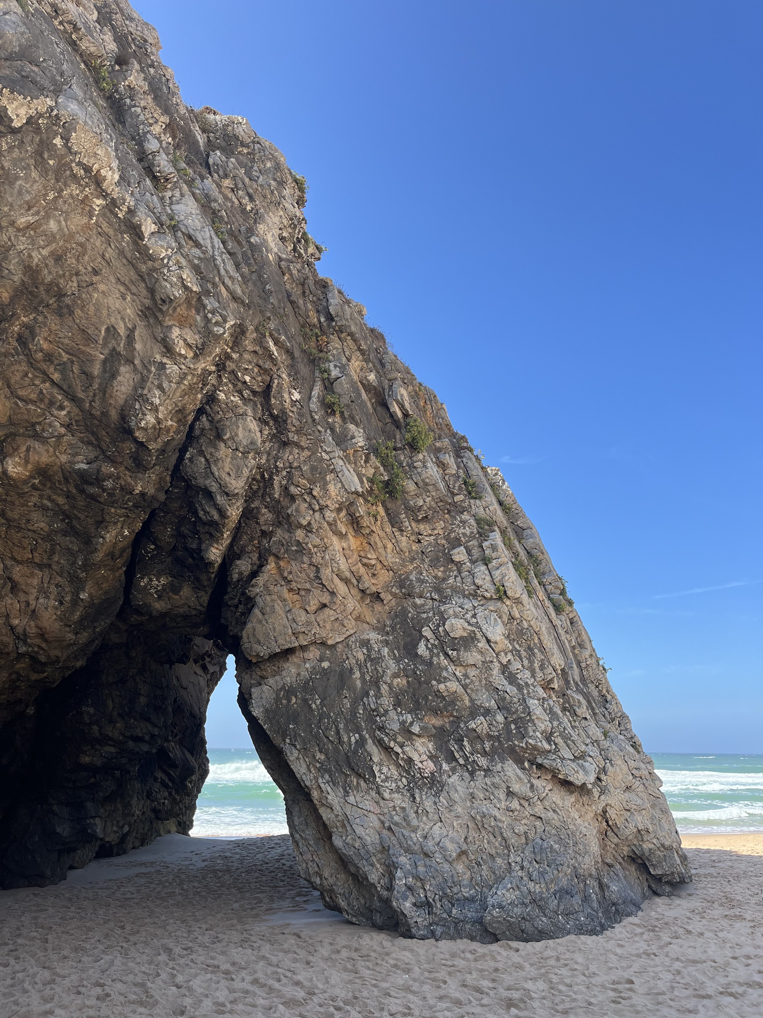 Large coastal rock formation with a natural arch and a small opening at the base, on a sandy beach with the ocean and blue sky in the background.