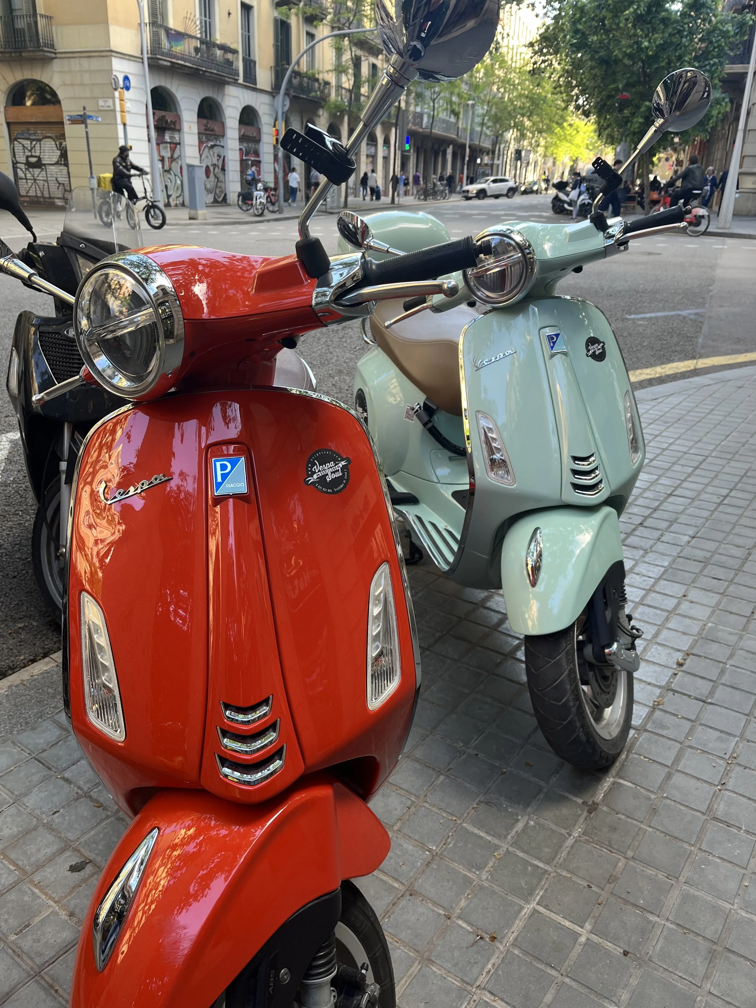 Two Vespa scooters parked on a city sidewalk, one red and one mint green, with buildings and pedestrians in the background.