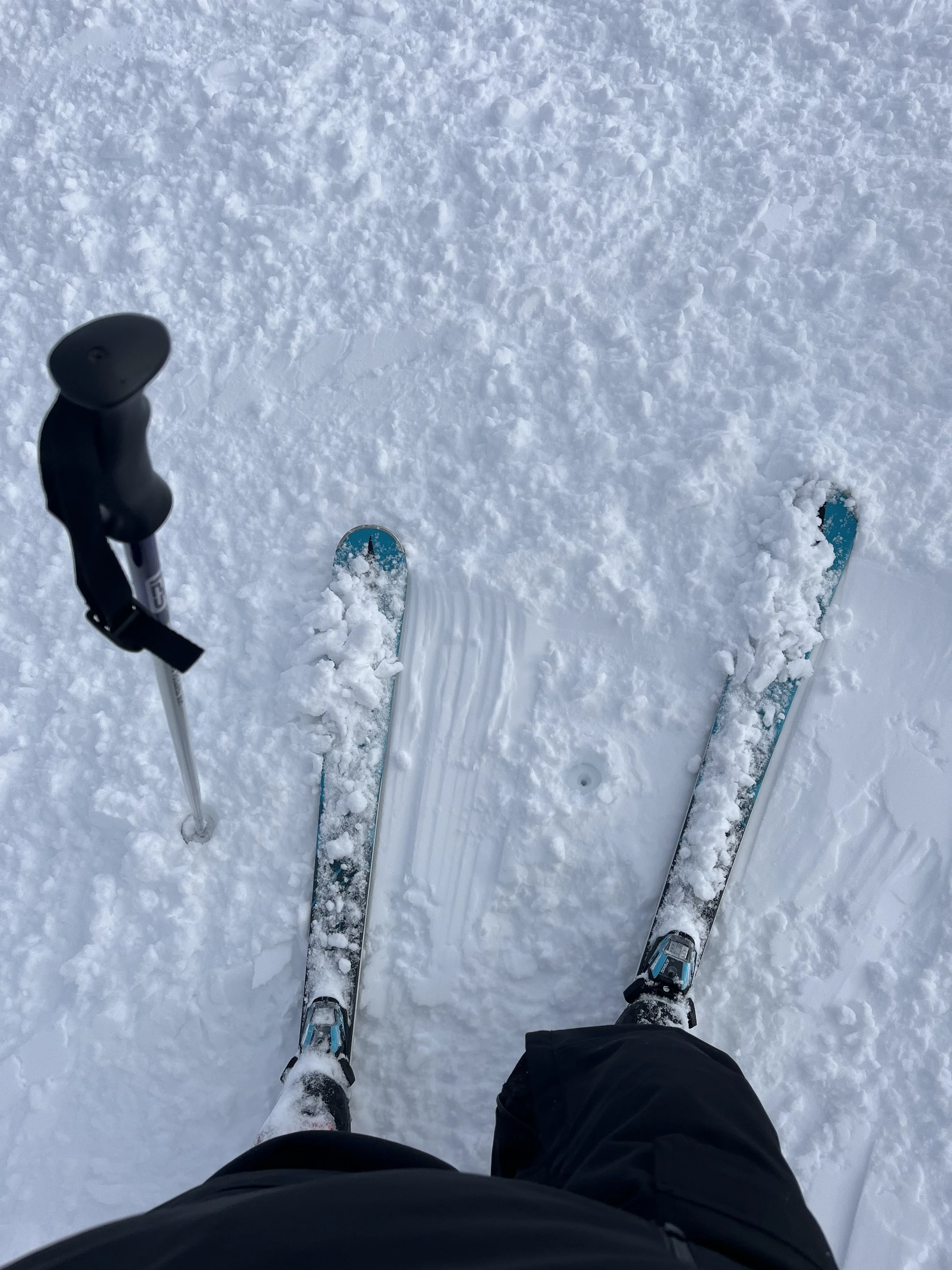 View from a person's perspective standing on skis on snow, with ski poles in hand; snow-covered ground appears groomed with some loose snow.