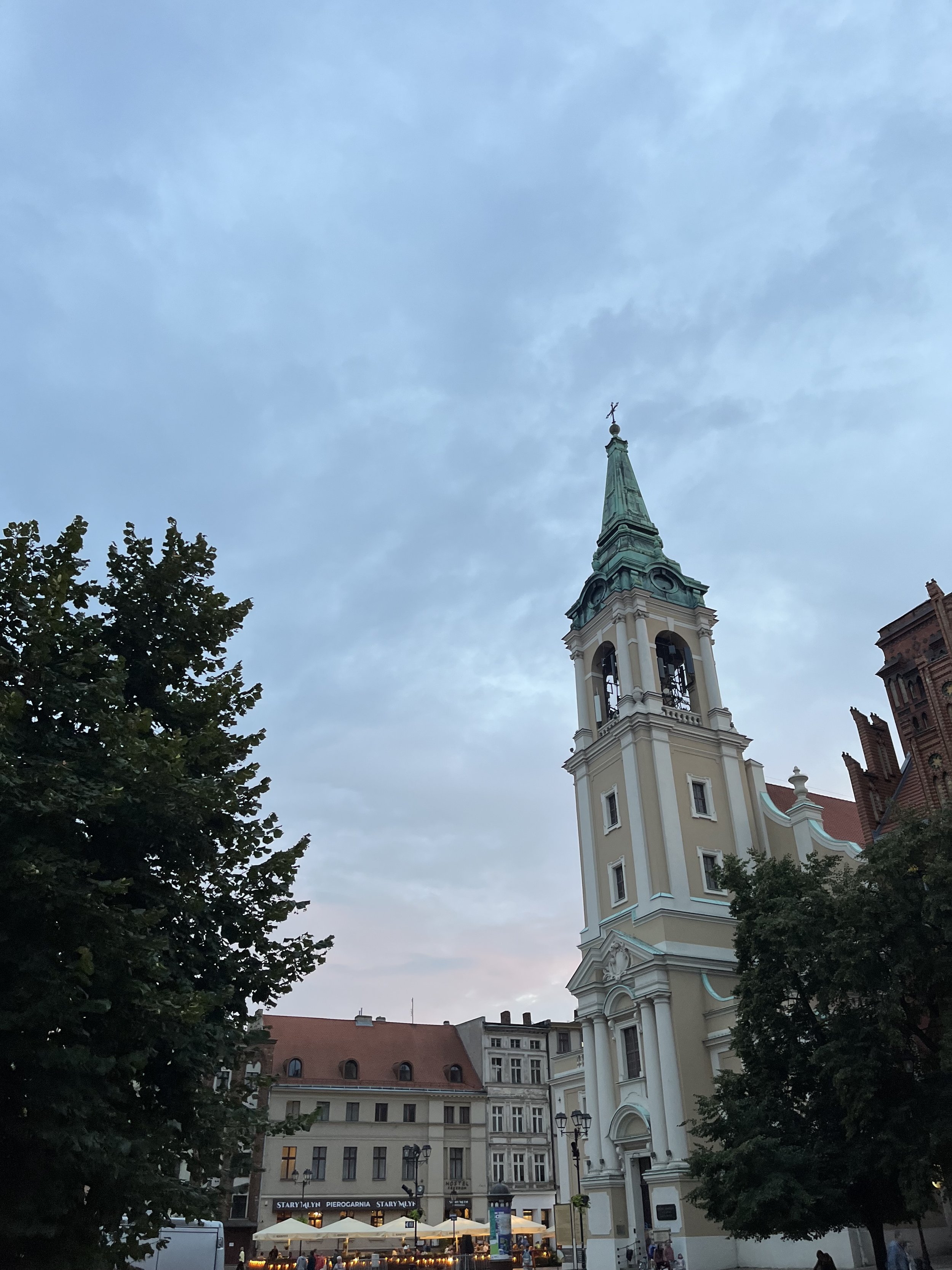 Tall church steeple with a cross on top in an urban square at dusk, with trees and historic buildings, and outdoor seating at a restaurant.