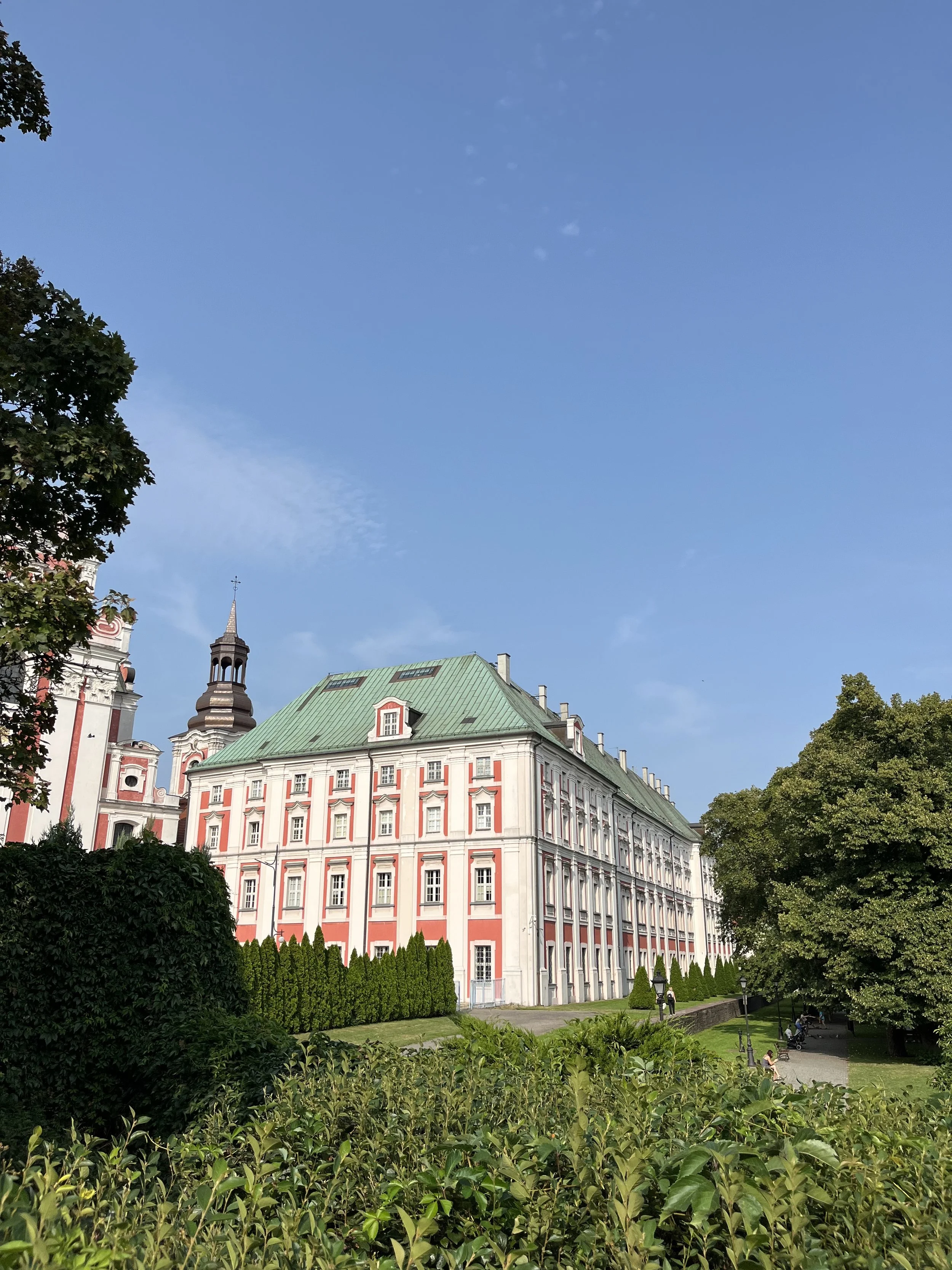 A historic building with a green roof and pink and white exterior, situated in a park with green trees and a clear blue sky.