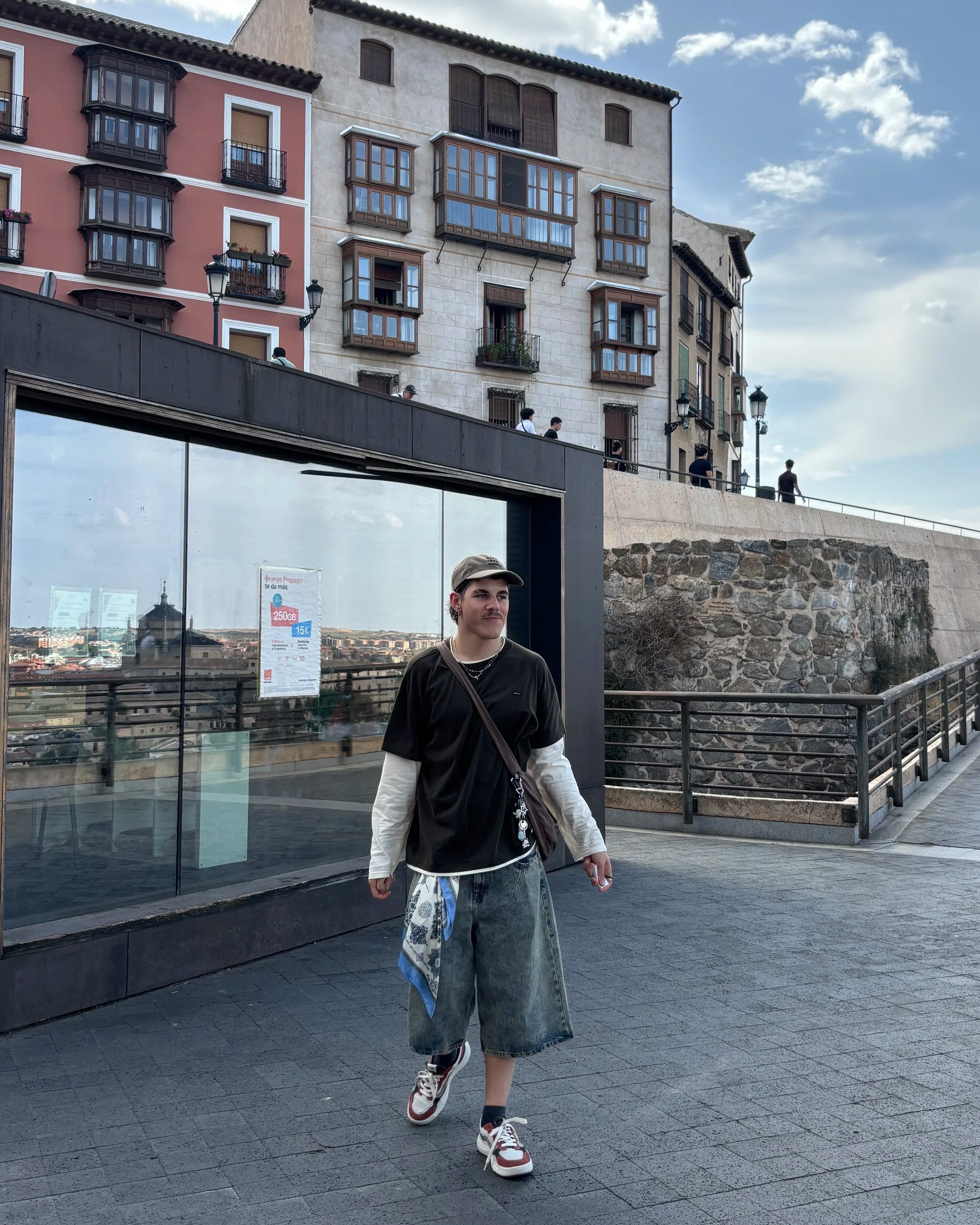 A young person walking along a street with a glass storefront behind them, behind which is a cityscape. The background features a row of buildings with balconies and a blue sky with clouds.