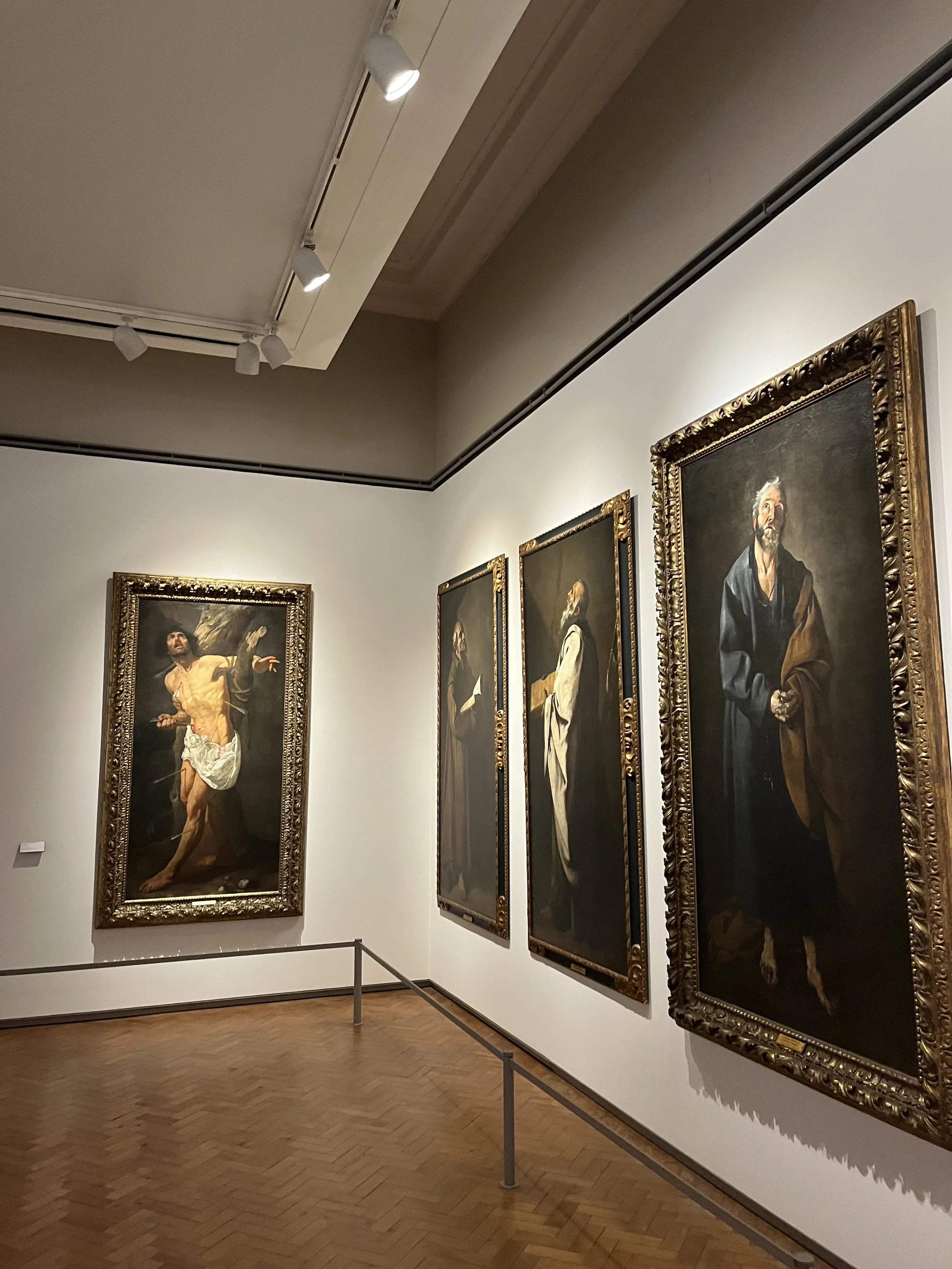 Gallery of paintings in a museum, featuring religious and historical figures in dark attire, with one depicting a man in white with a beard, and another showing a man with a dark beard and a brown cloak.