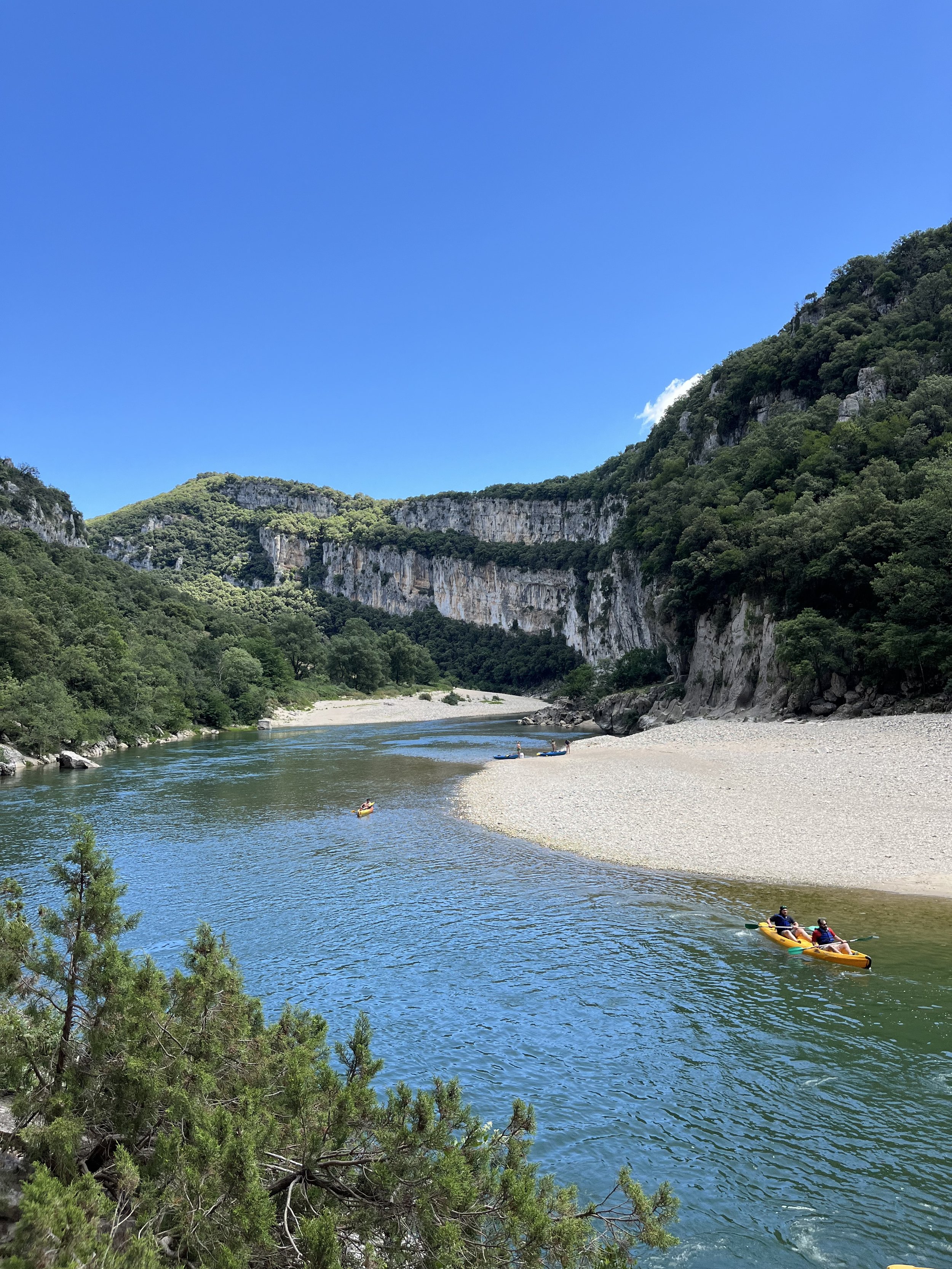 People kayaking on a calm river surrounded by lush green trees and steep cliffs under a clear blue sky.