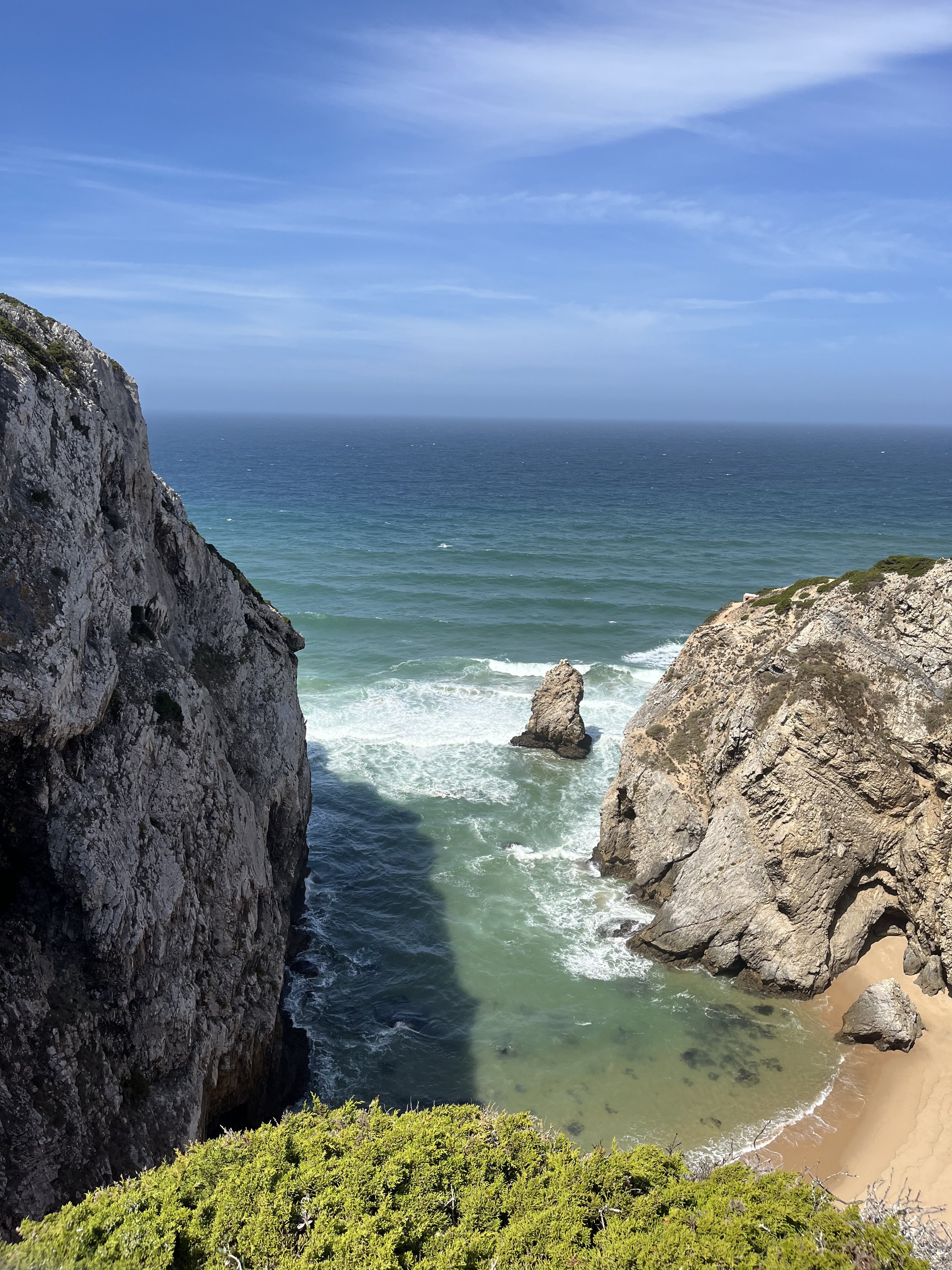 A scenic view of a beach with cliffs on either side, a large rock in the water, and a sandy shore at the bottom right.
