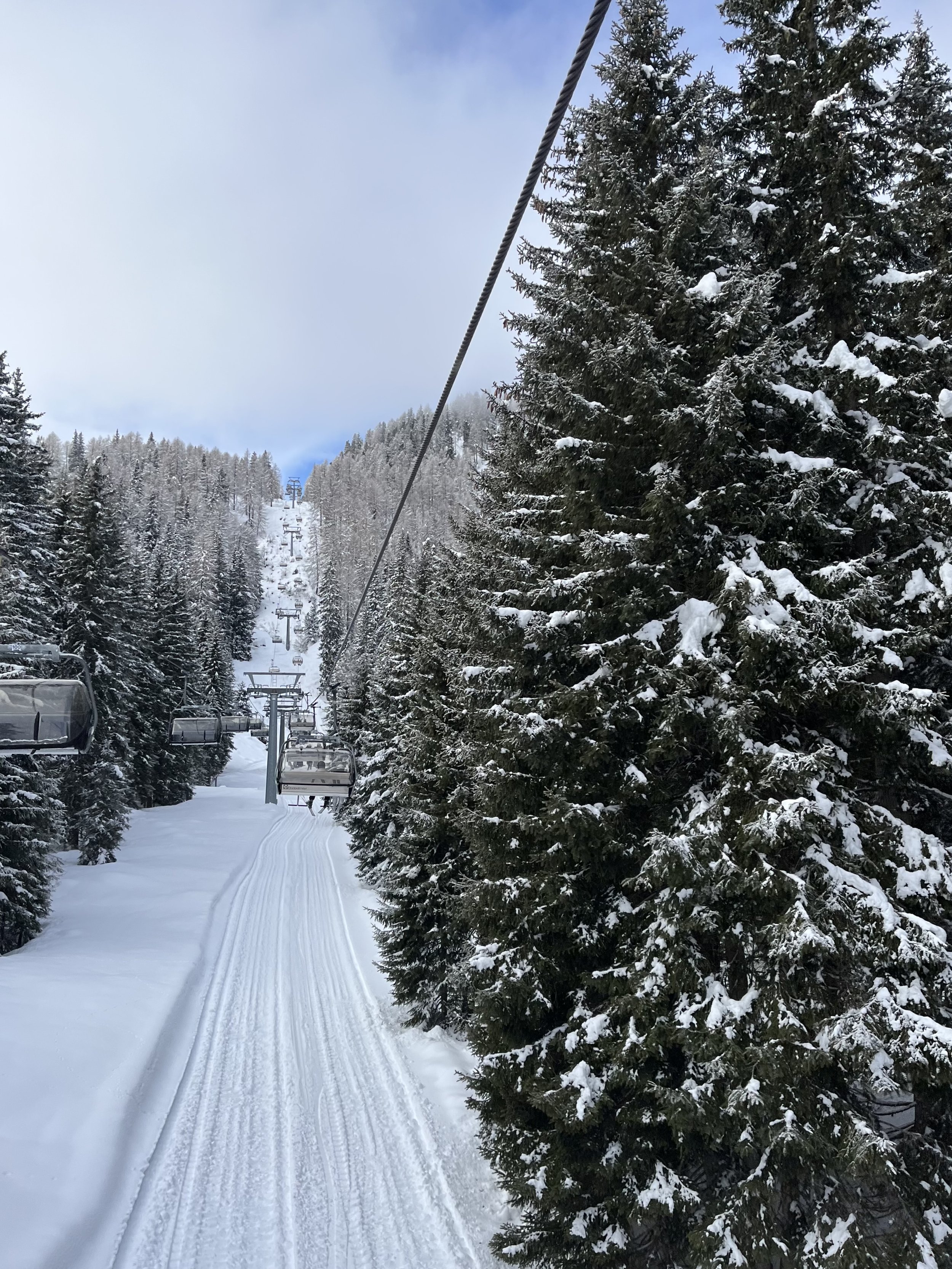 Ski lift ascending a snow-covered mountain with trees on either side and a trail of ski tracks below, under a cloudy sky.