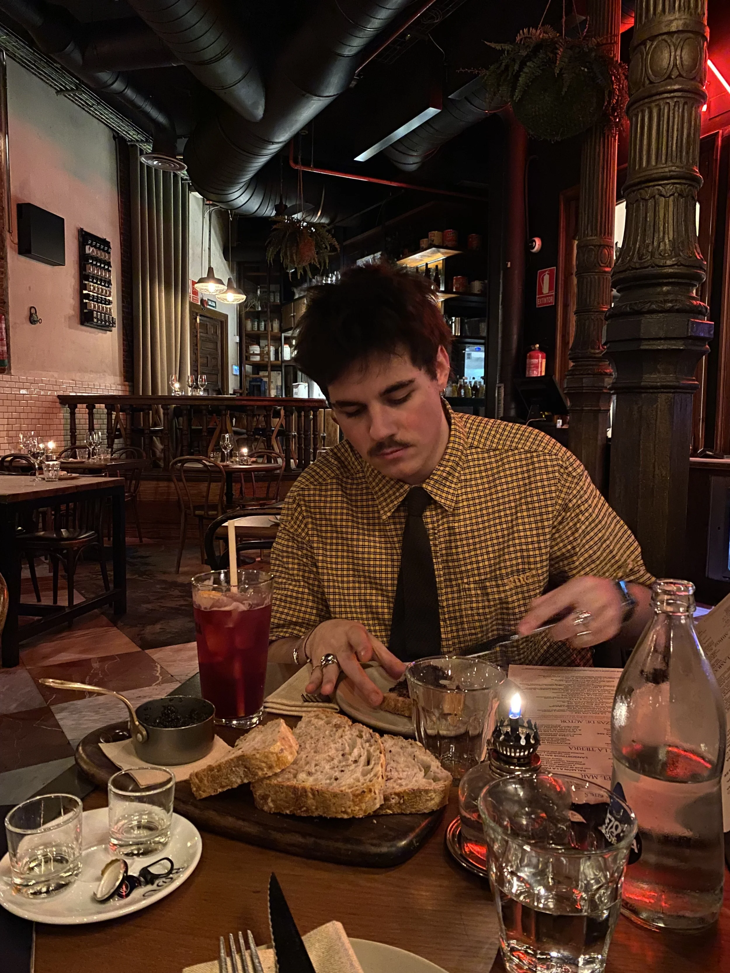 A young man with dark hair, a mustache, and light skin, wearing a yellow checkered shirt and black tie, is seated at a restaurant table, cutting into a plate of food. The table has slices of bread, a glass of dark red beverage with a straw, and various drinks and utensils. The restaurant interior has warm dim lighting, wooden furniture, and decorative suspending plants.